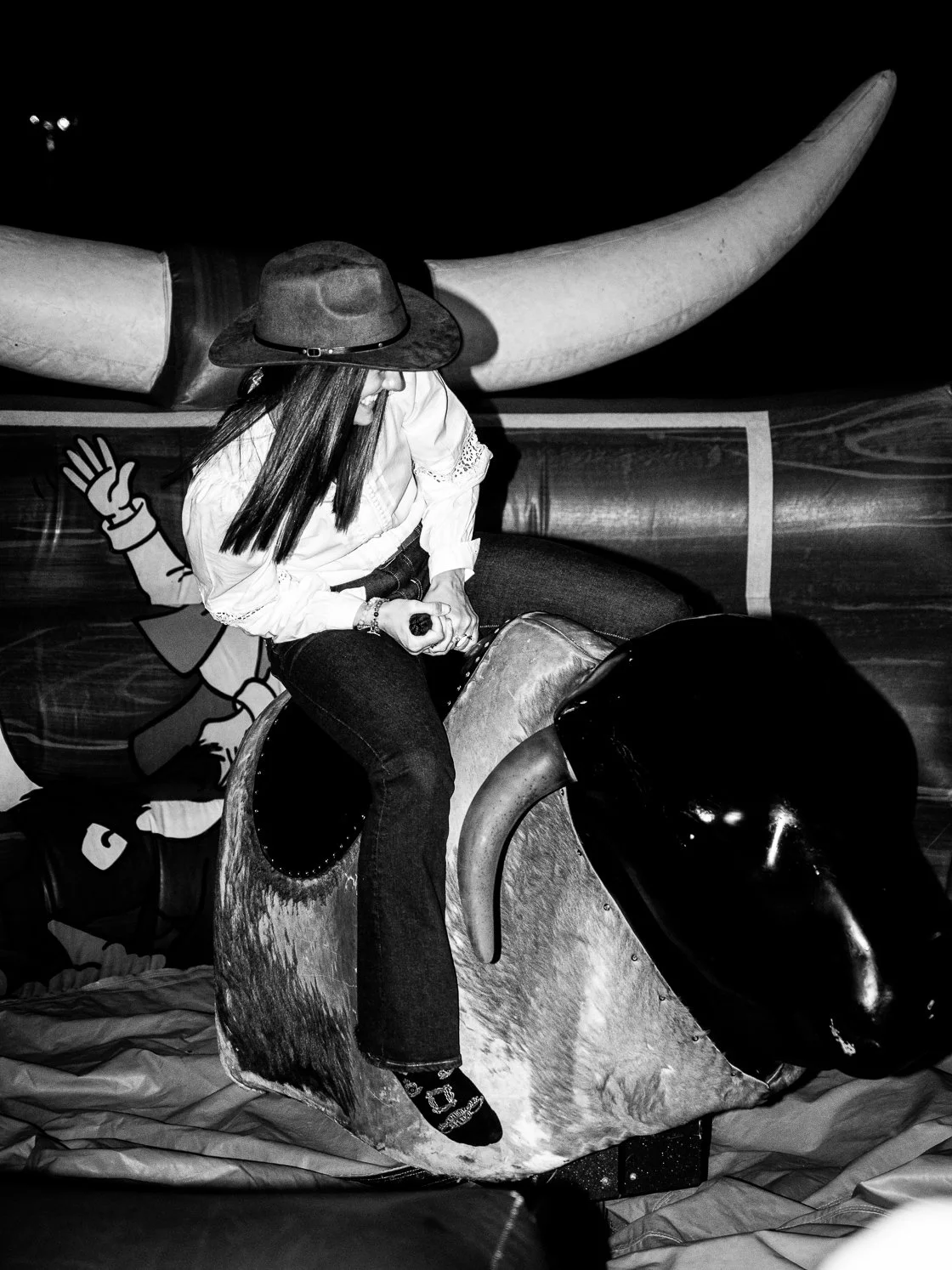A woman wearing a hat and white shirt riding a mechanical bull at an indoor amusement venue.