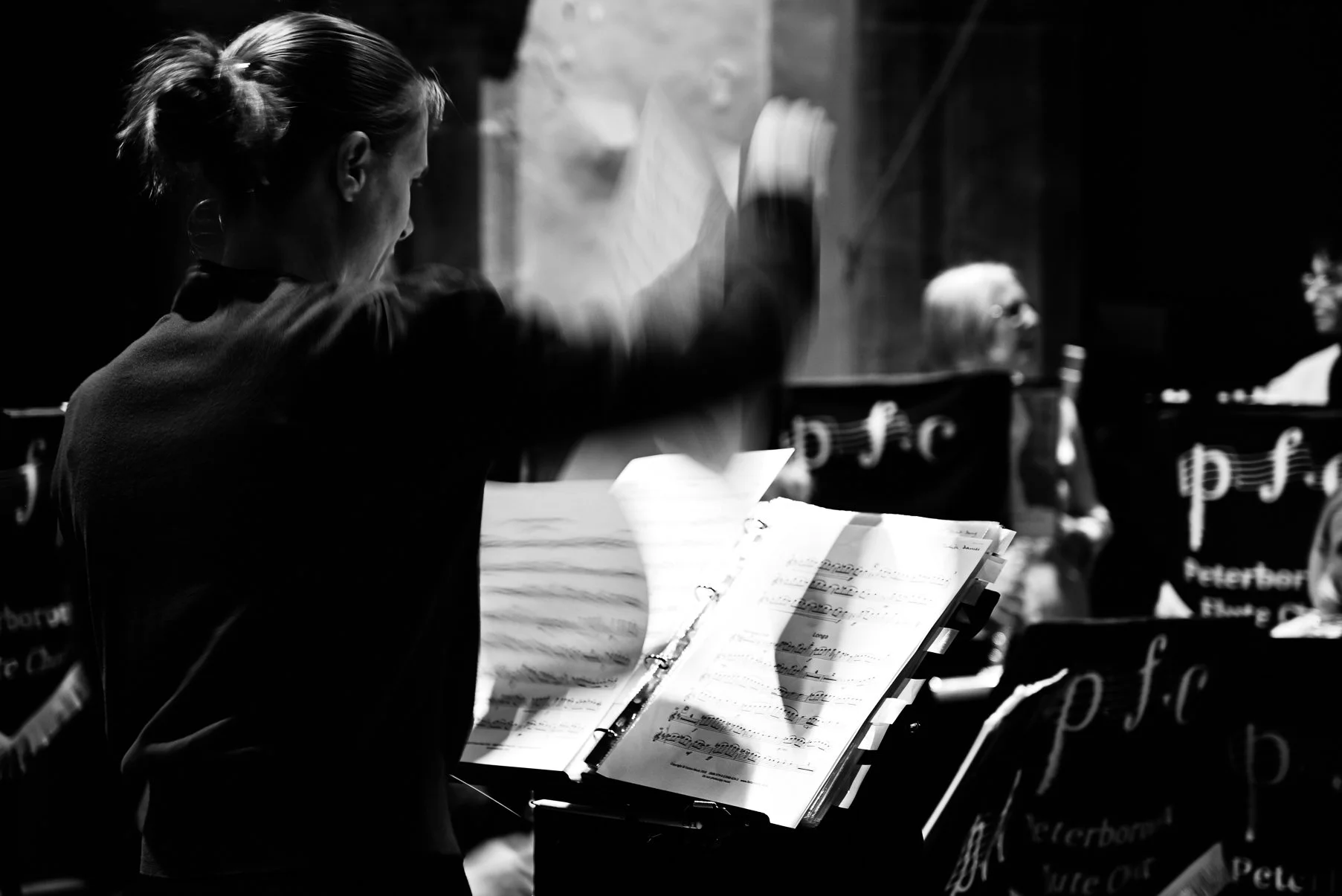 A black and white photo of a woman conducting an orchestra, viewed from the side. She is focusing on sheet music on a music stand, with other musicians and music stands visible in the background.