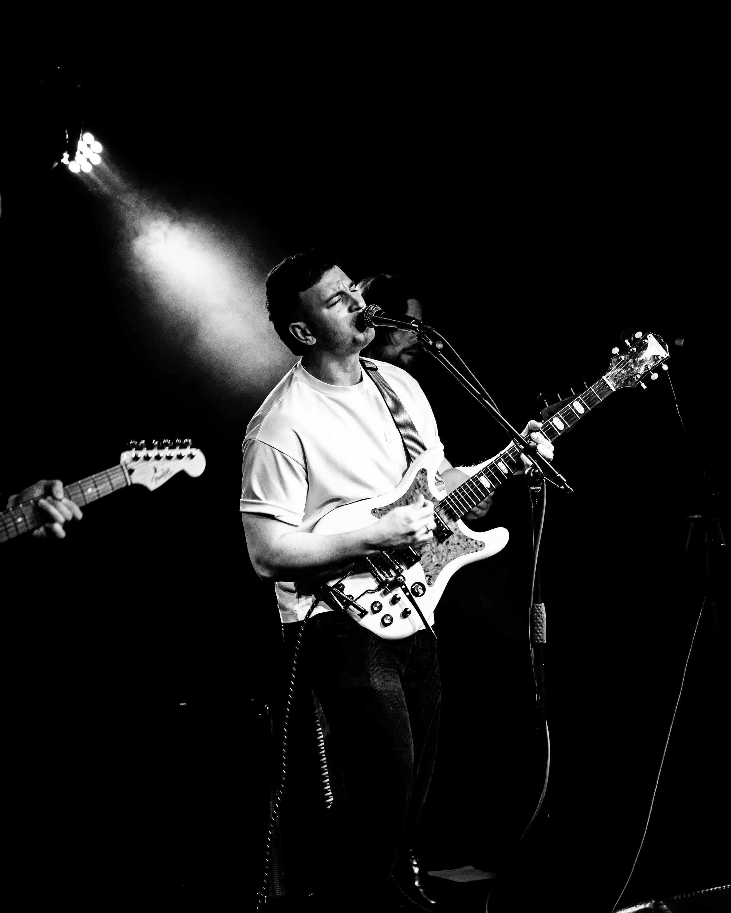 A black and white photograph of a young man singing into a microphone while playing an electric guitar on stage.