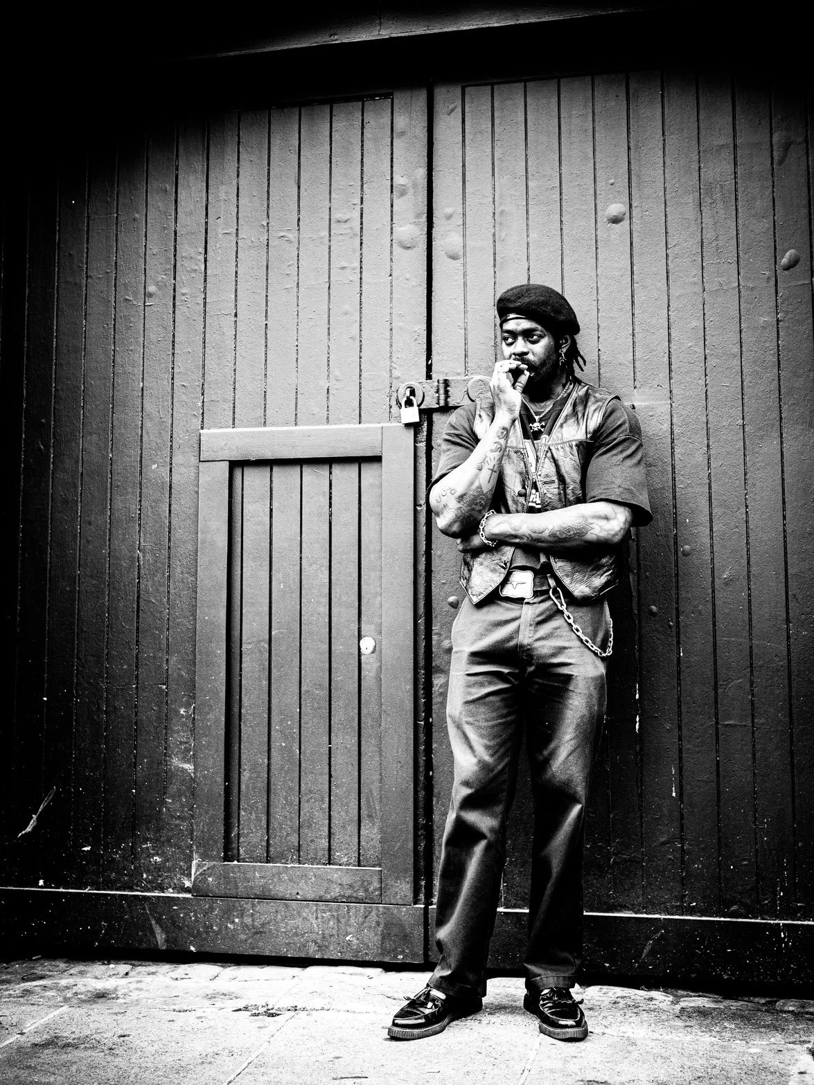 A man standing in front of a large wooden door, wearing a beret, leather vest, dark pants with a chain, tattoos on his arms, and smoking a cigarette.