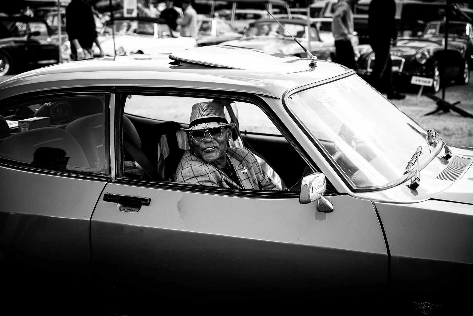 A man wearing sunglasses, a hat, and a plaid jacket is sitting in the driver's seat of a classic sports car at an outdoor car show. The background features several other vintage cars and people walking around.