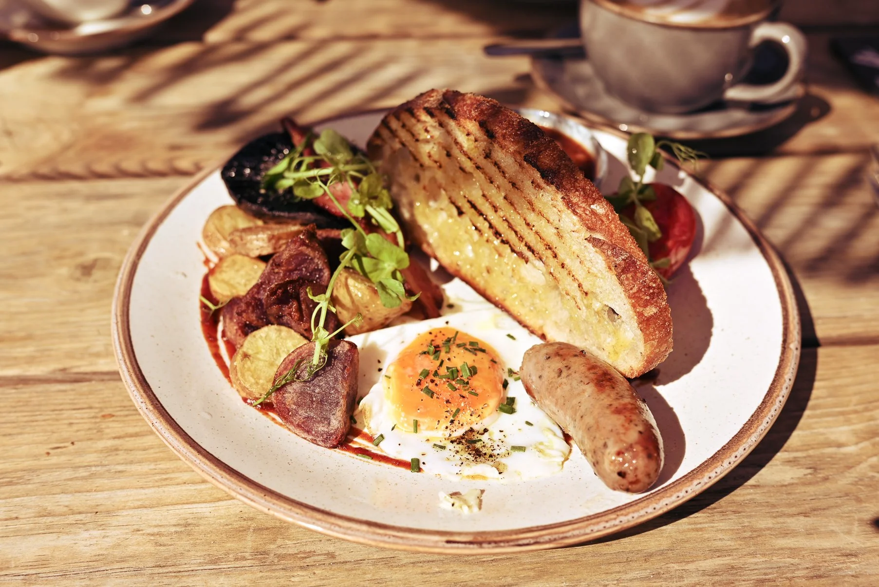 A plate of breakfast food with toasted bread, fried egg, sausage link, roast potatoes, and sausage pieces, garnished with microgreens, on a wooden table.
