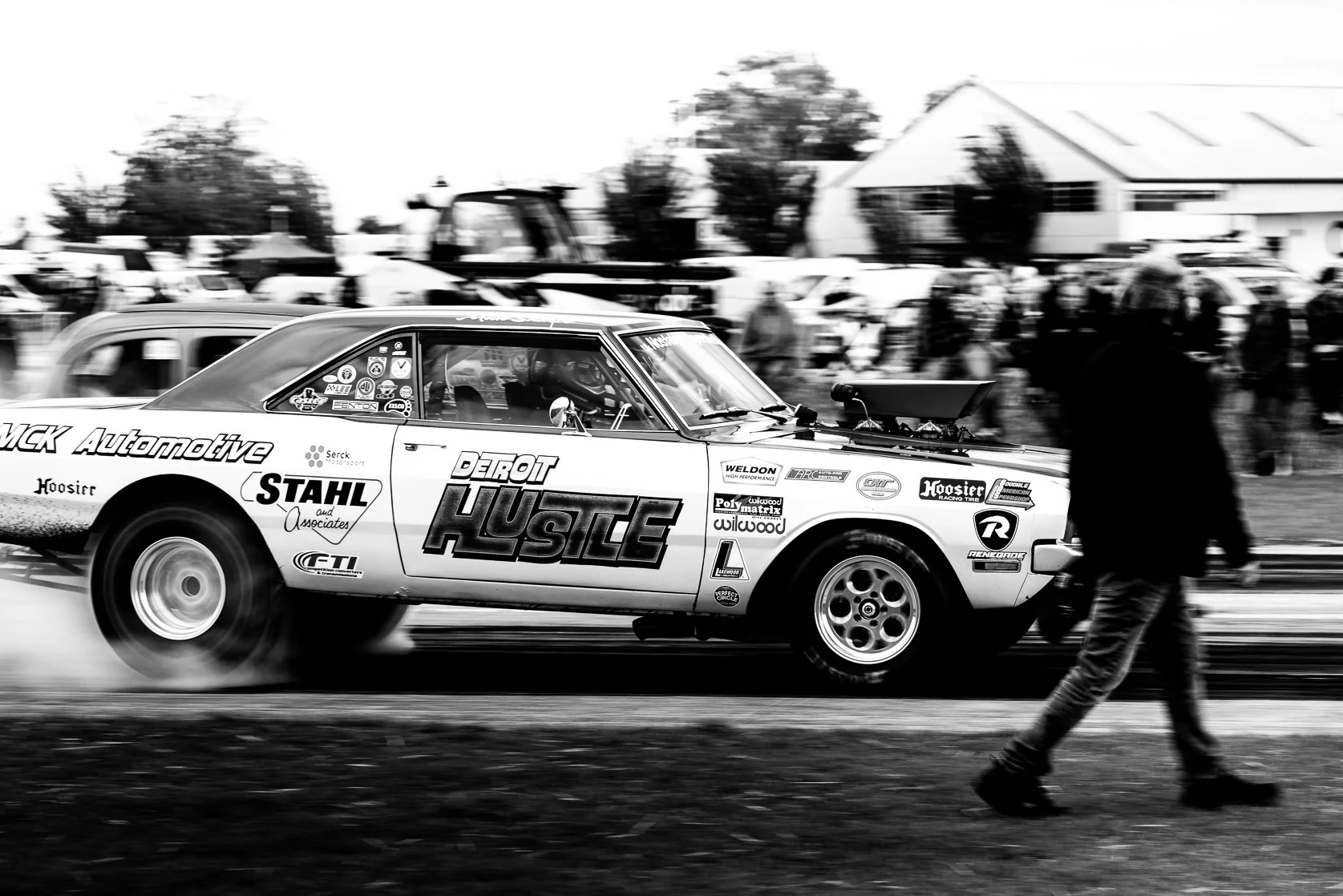 A black and white race car with sponsorship stickers and the text 'Detroit Hustle' is racing on a track, with a person walking in the foreground, and spectators and buildings in the background.