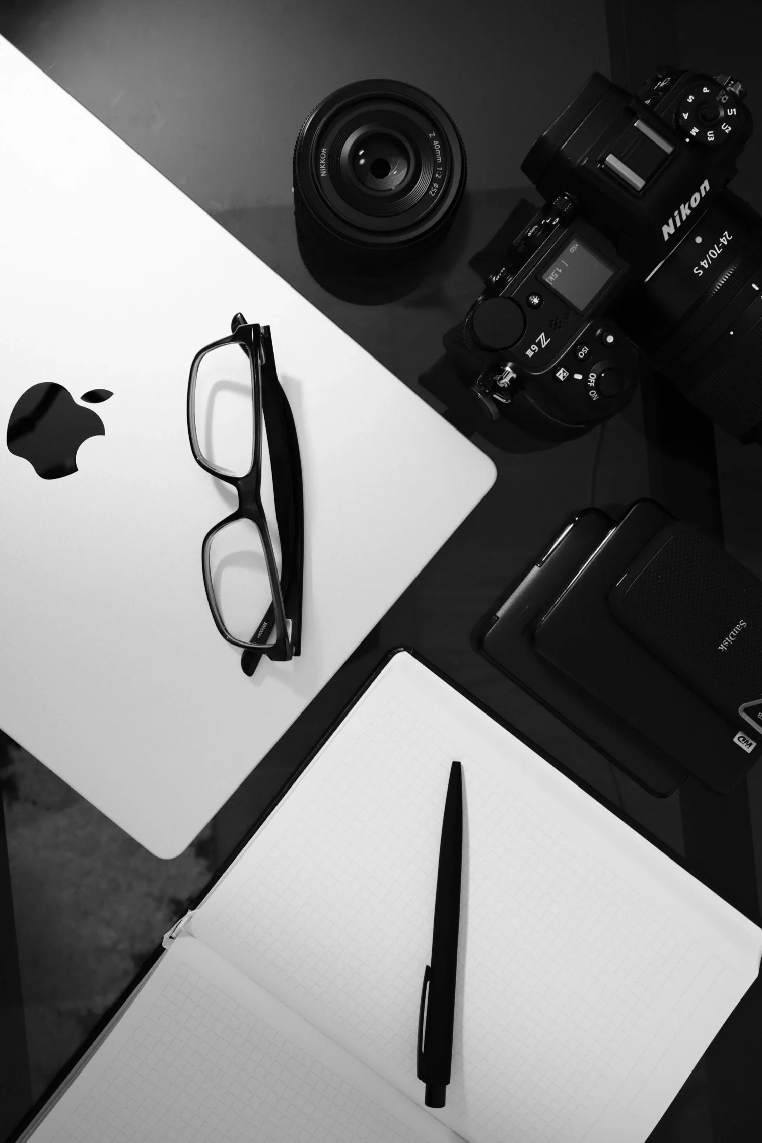 Black and white photo showing a computer with glasses, a camera, a notepad with grid paper, and a pen on a desk with two-tone surface.