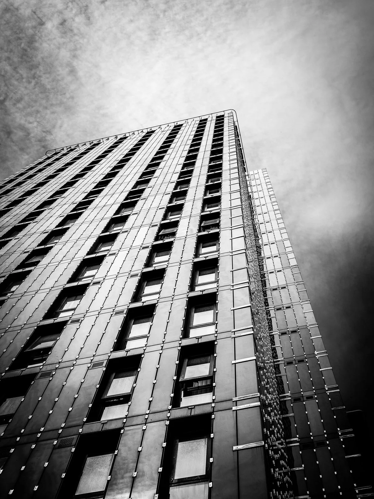 Black and white photo of a tall modern building with grid-like windows and metallic facade, taken from a low angle against a cloudy sky.