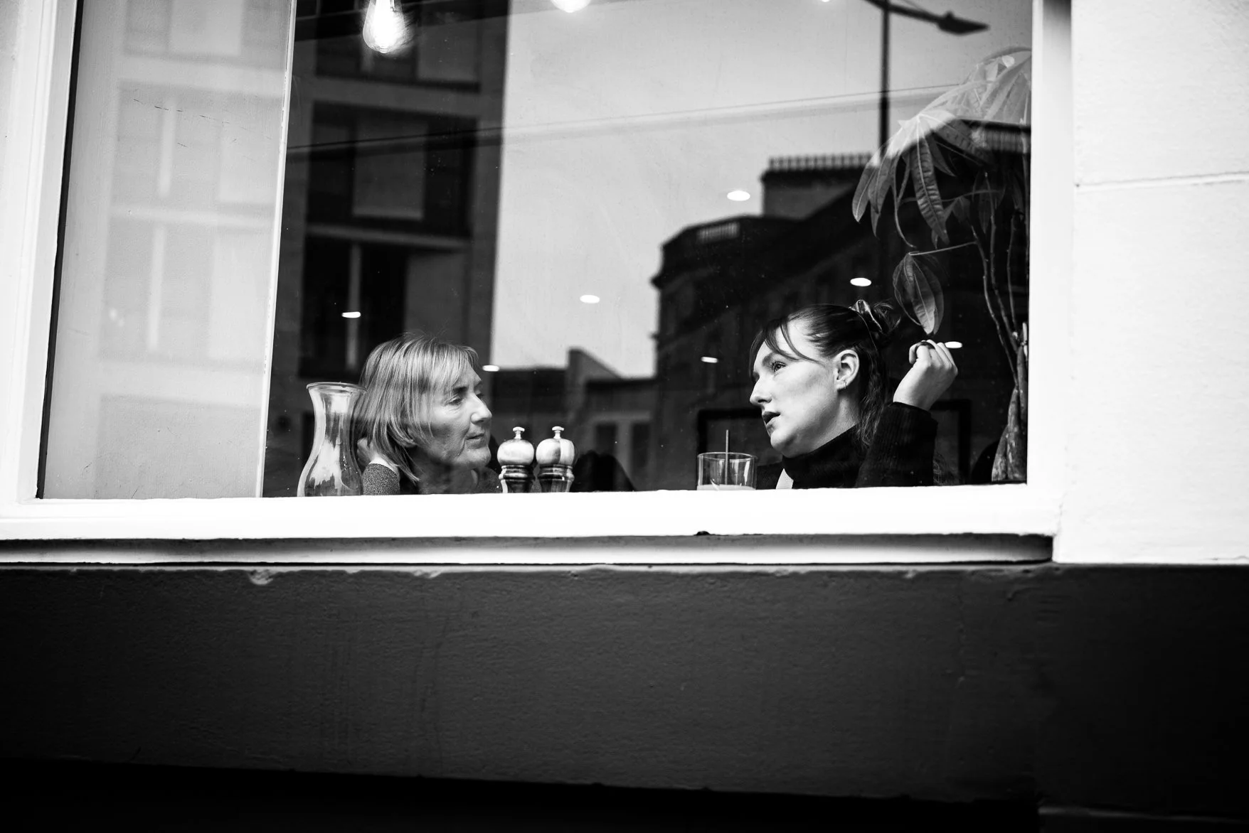 View through a window showing two women sitting inside a restaurant or cafe, engaged in conversation, with a glass and a pitcher on the table, and a plant in the background.