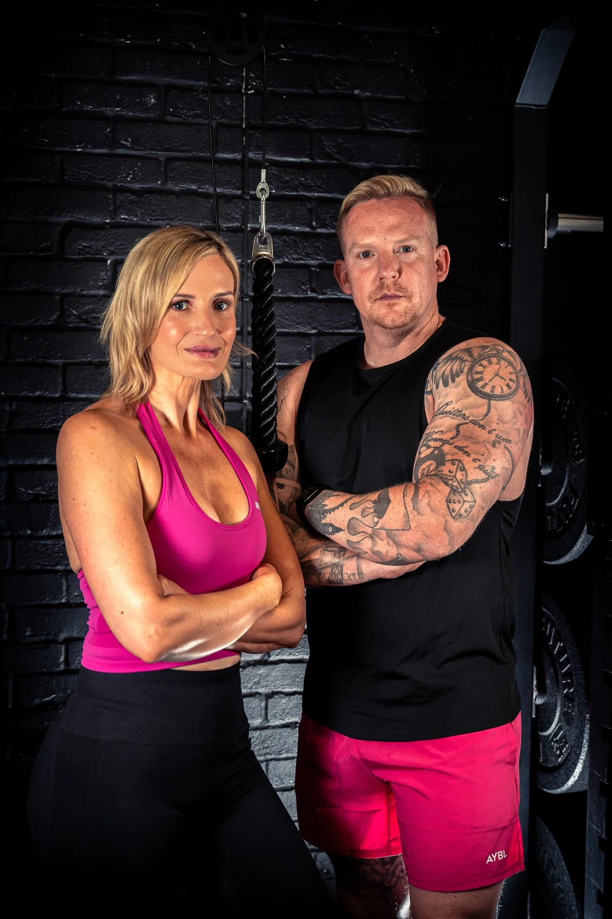 A woman in a pink sports bra and black leggings standing with arms crossed; a man with tattoos in a black sleeveless shirt and red shorts, with arms crossed, posing in a gym with black brick wall and weightlifting equipment in the background.