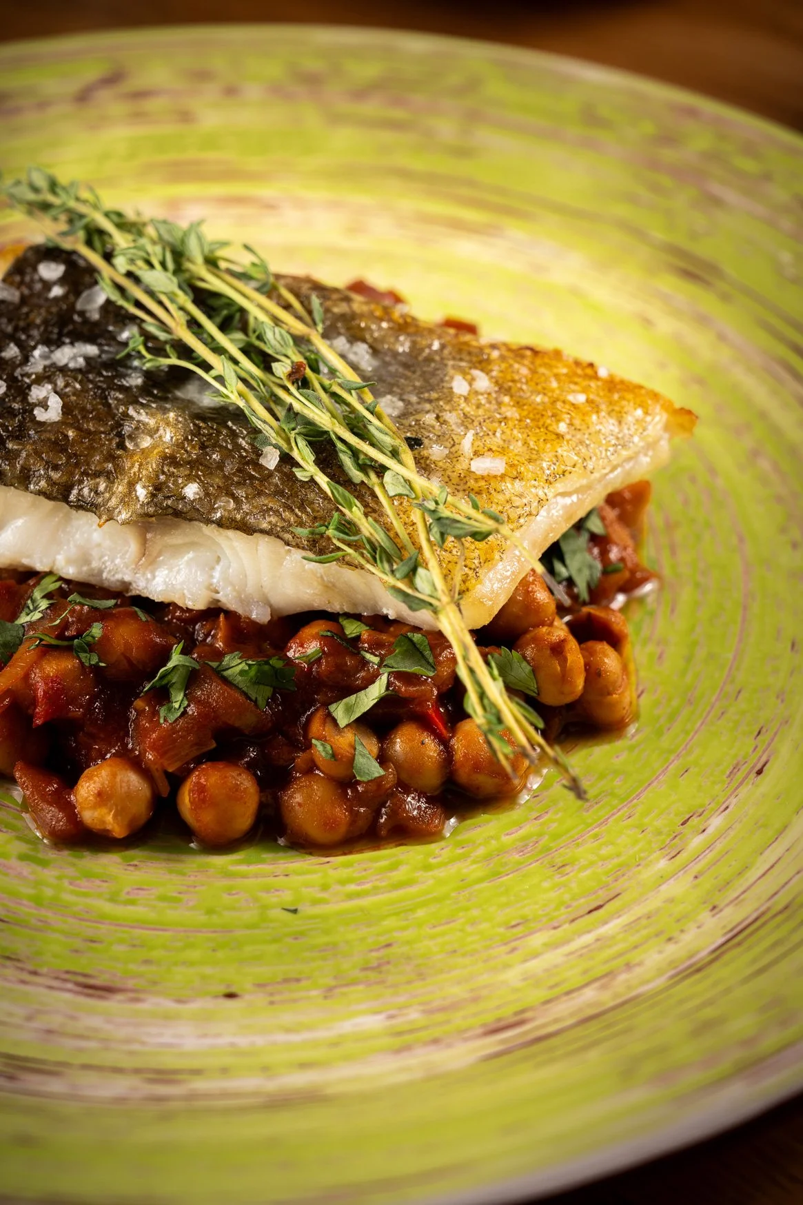 A plated dish featuring cooked chickpeas in a tomato-based sauce, topped with an seared fish fillet, garnished with fresh thyme sprigs and coarse salt.