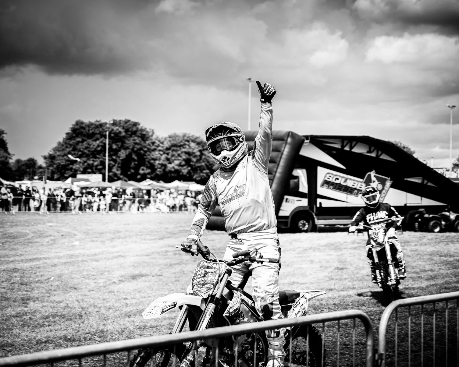 Black and white photo of a motocross rider on a motorcycle raising one hand in the air during a race event. There is a crowd of spectators in the background along with tents and a large inflatable arch with the words 'Soubb FSLER' visible, and a seco