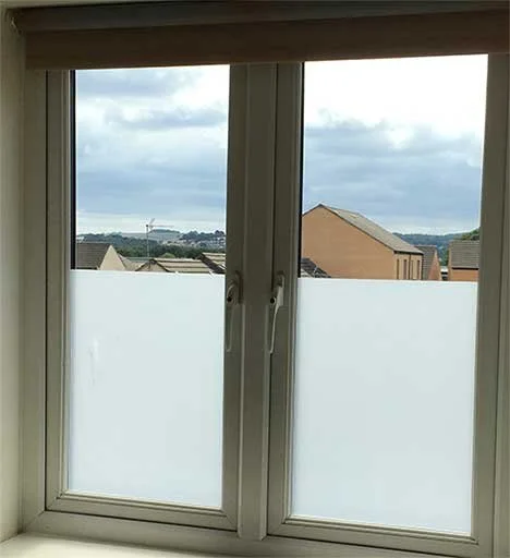 View of rooftops and distant buildings through a double-pane window with frosted lower panels, under a partly cloudy sky.