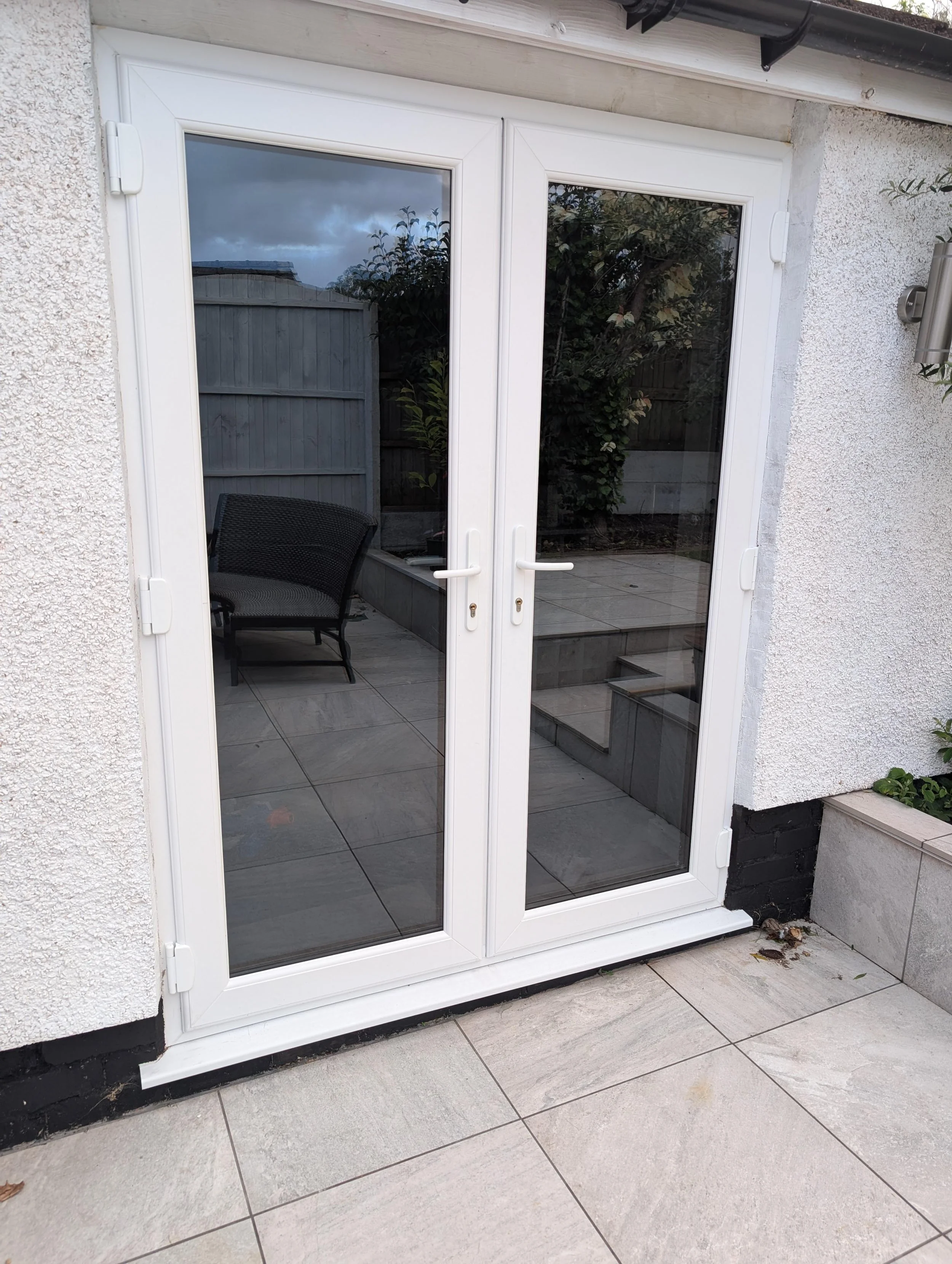 A set of white double glass patio doors leading to an outdoor patio area with gray tiled flooring and a black patio chair. The doors are framed by a textured white wall, with a small outdoor wall-mounted light fixture on the right side. There are some plants and a wooden fence visible outside.