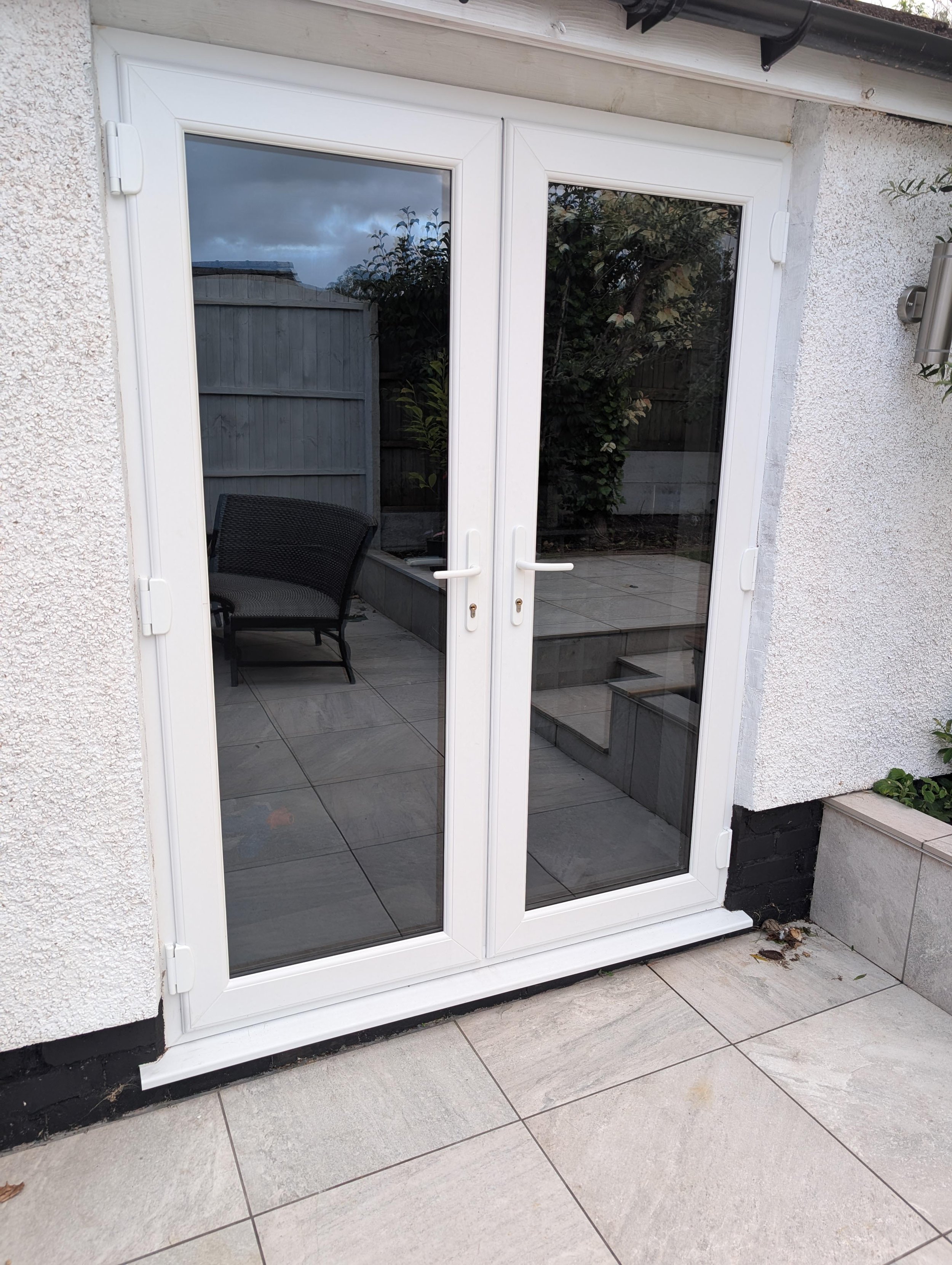 A set of glass double doors with white frames leading outside. The doors reflect the cloudy sky and some trees. The exterior walls are textured white, and there are tiled steps and patio outside.