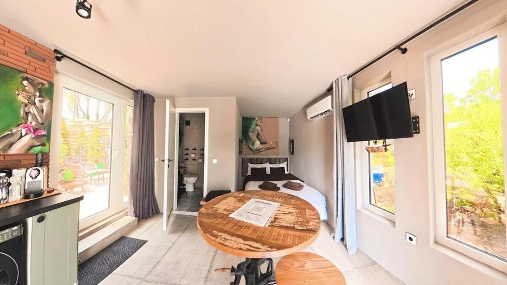 Bright, cozy bedroom with a double bed, mounted TV, small bathroom, and glass door leading outside, with a round wooden table in the foreground.