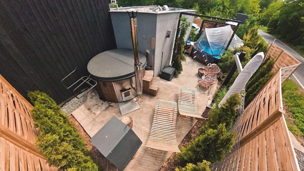 A rooftop outdoor patio with a hot tub, wooden chaise lounges, and various chairs, surrounded by green trees and enclosed by a wooden fence.