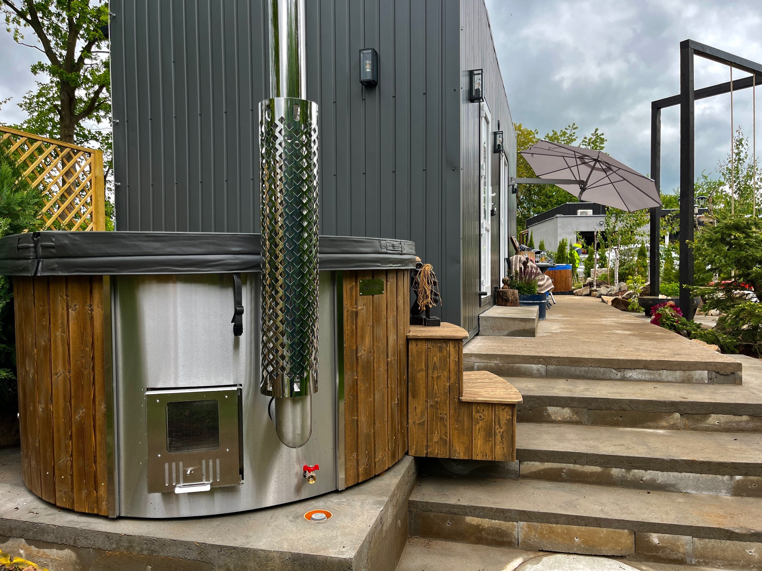 An outdoor space with a container hot tub on a concrete pad, a stainless steel outdoor heater, a wooden staircase leading to a deck, a gray wall with black outdoor lights, a large umbrella, and various plants and trees under a cloudy sky.
