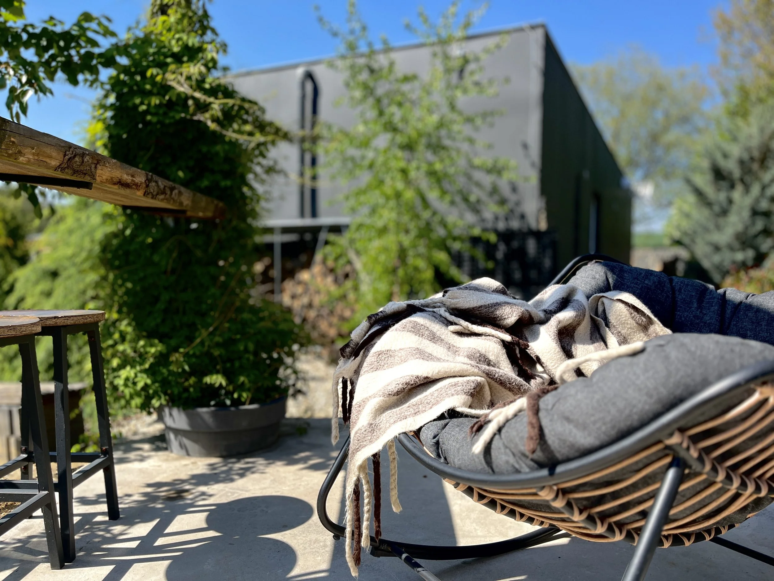 A wicker chair with a blanket draped over it, situated on a porch or patio. In the background, a large gray house with black accents is visible, surrounded by trees and greenery under a clear blue sky.