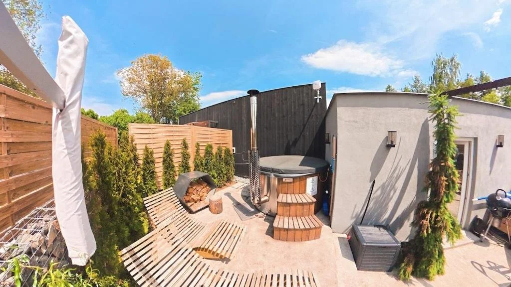 Backyard with a hot tub, wooden stairs, and outdoor seating area. Tall evergreen shrubs line a wooden fence, and a tall, narrow evergreen tree with a shadow on the wall. Clear blue sky with a few clouds.