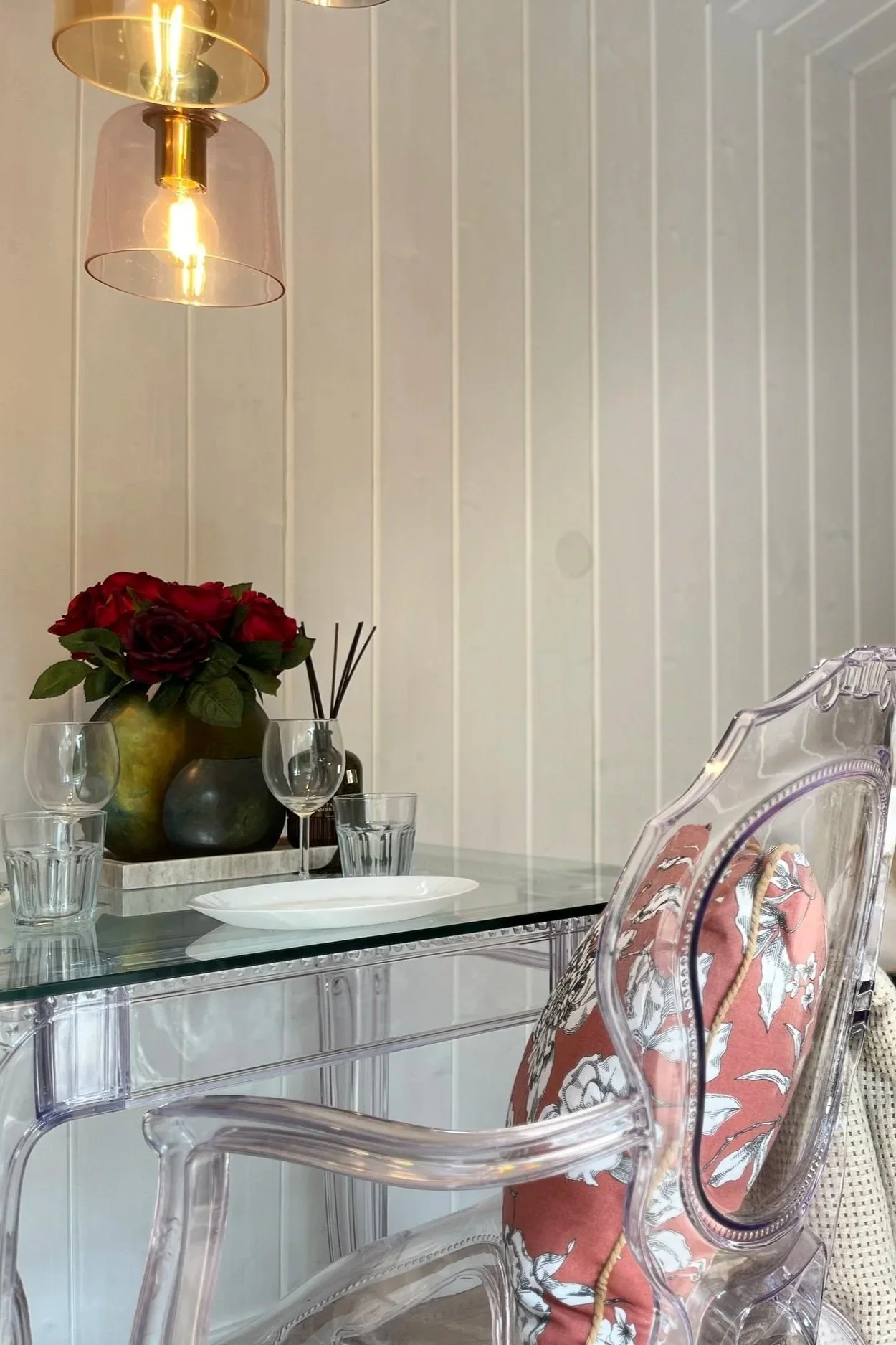 A dining room with a glass table set with four glasses, a white oval plate, and a centerpiece with red roses in a green and black vase, set against a white paneled wall illuminated by two pendant lights with pink and gold shades.
