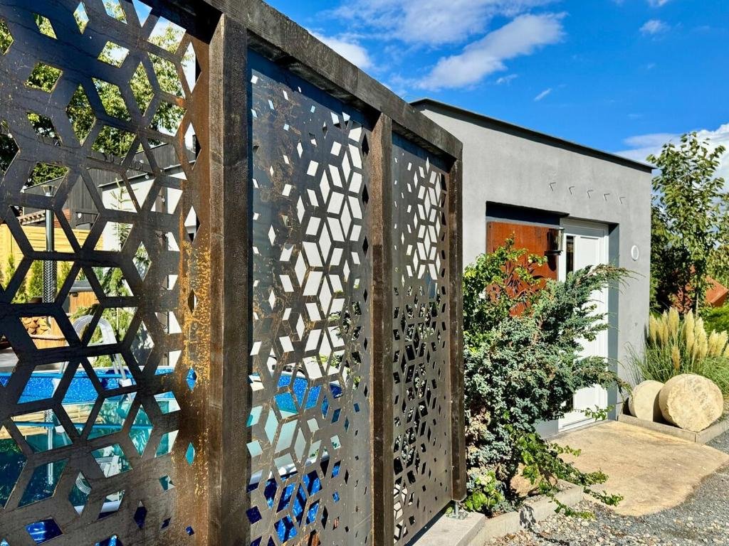 Modern house with gray exterior walls, white sliding door, and lush green shrubs in the front yard. Decorative metal gate with geometric pattern in the foreground, blue sky with clouds above.