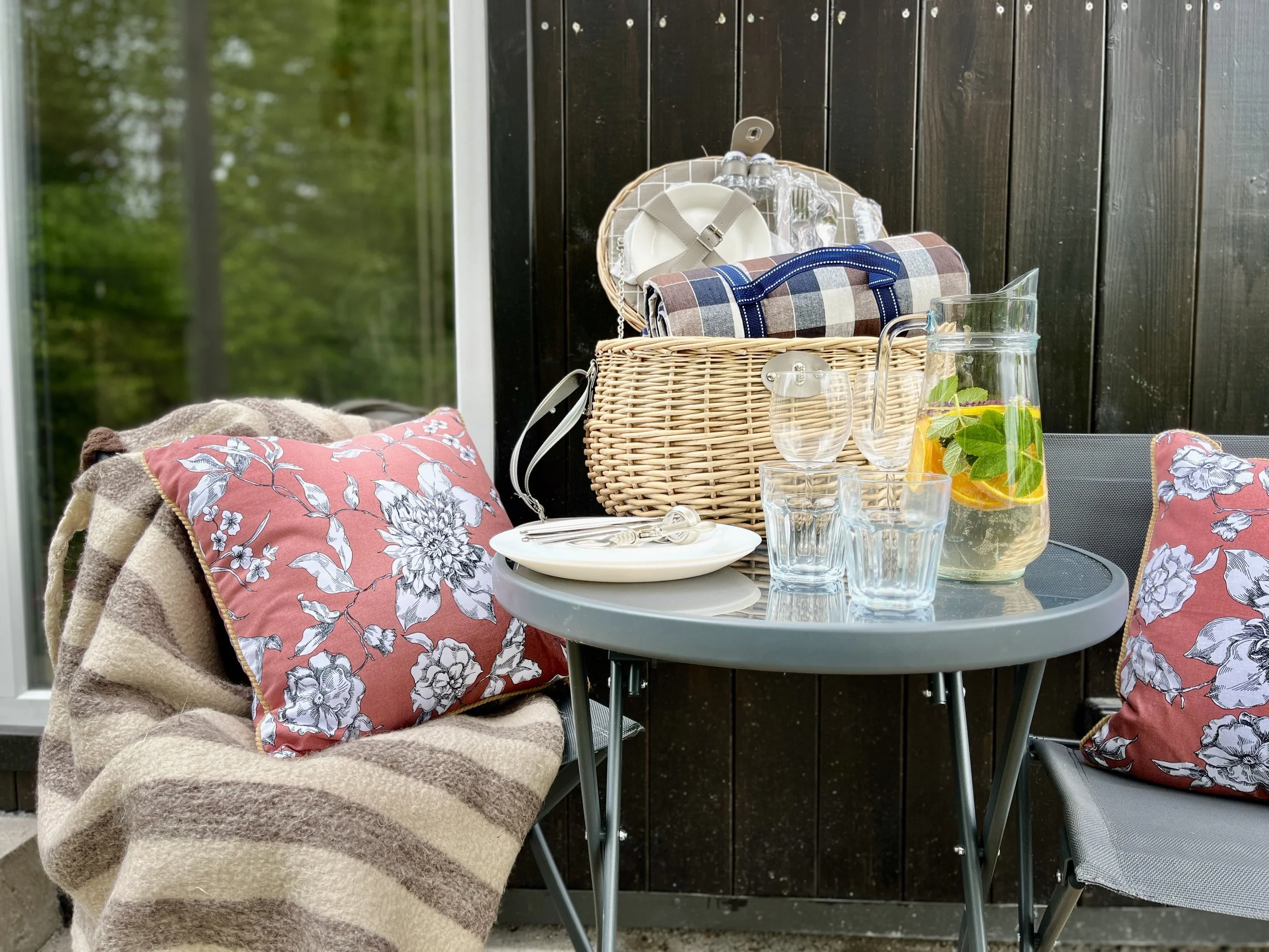 A round patio table with a pitcher of lemon mint infused water, several empty glasses, and a plate with cutlery on it. Two chairs are visible, one with a floral cushion and soft blanket, the other with a floral cushion. There is a wicker picnic basket filled with plates, a cloth napkin, and plastic utensils. The background features a dark wood fence and trees.