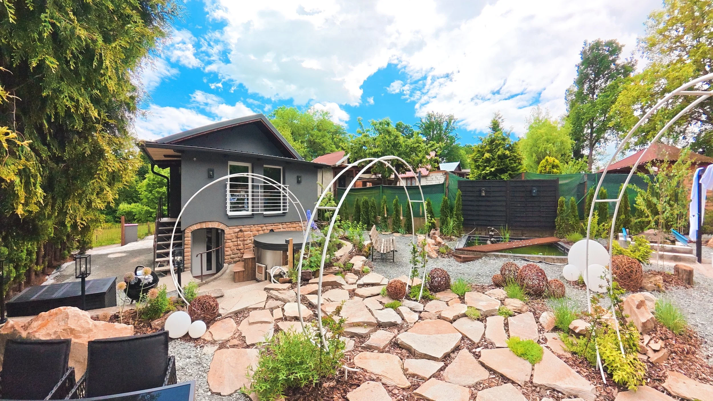 A backyard garden with stone paths, decorative garden spheres, a pond, and outdoor seating. There is a small house with modern windows and a sloped roof, surrounded by lush green trees and a bright blue sky with scattered clouds.