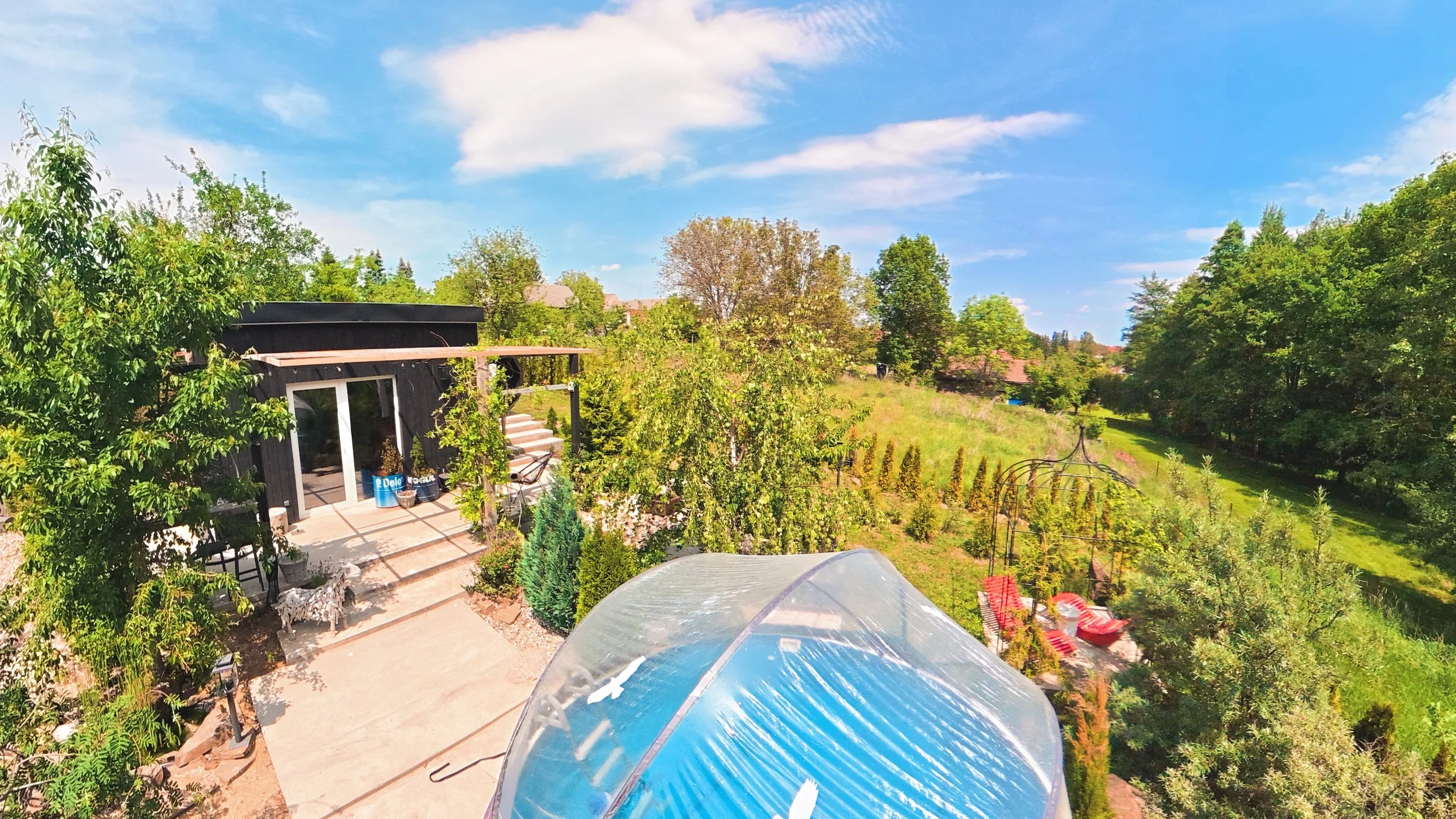 A backyard garden with trees, a patio with chairs, and a greenhouse in the foreground. The yard is surrounded by greenery and distant houses under a bright blue sky with some clouds.