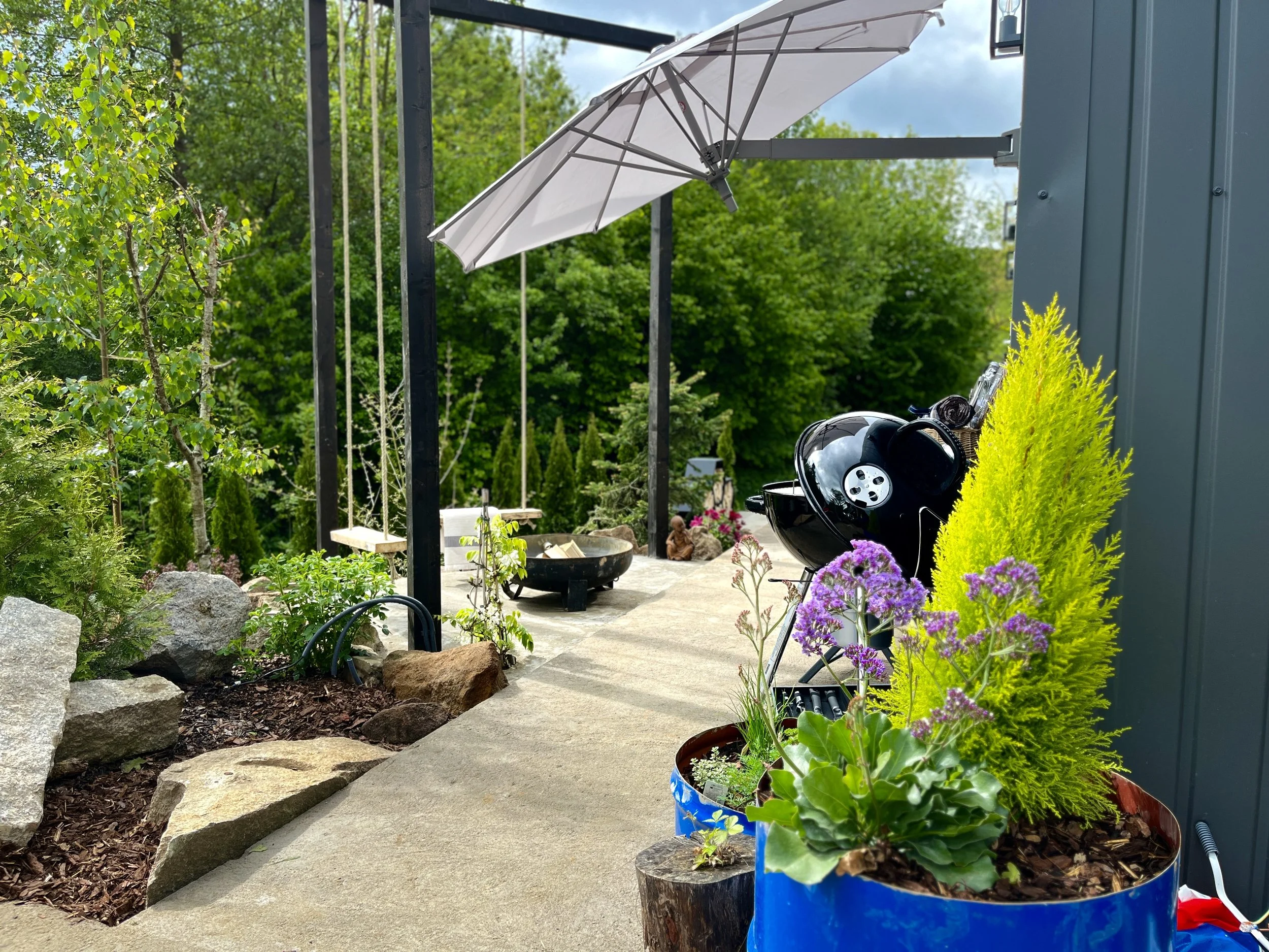 A backyard patio scene with potted plants, flowers, rocks, and a barbecue grill. A white patio umbrella is open. Green trees and blue sky are in the background.