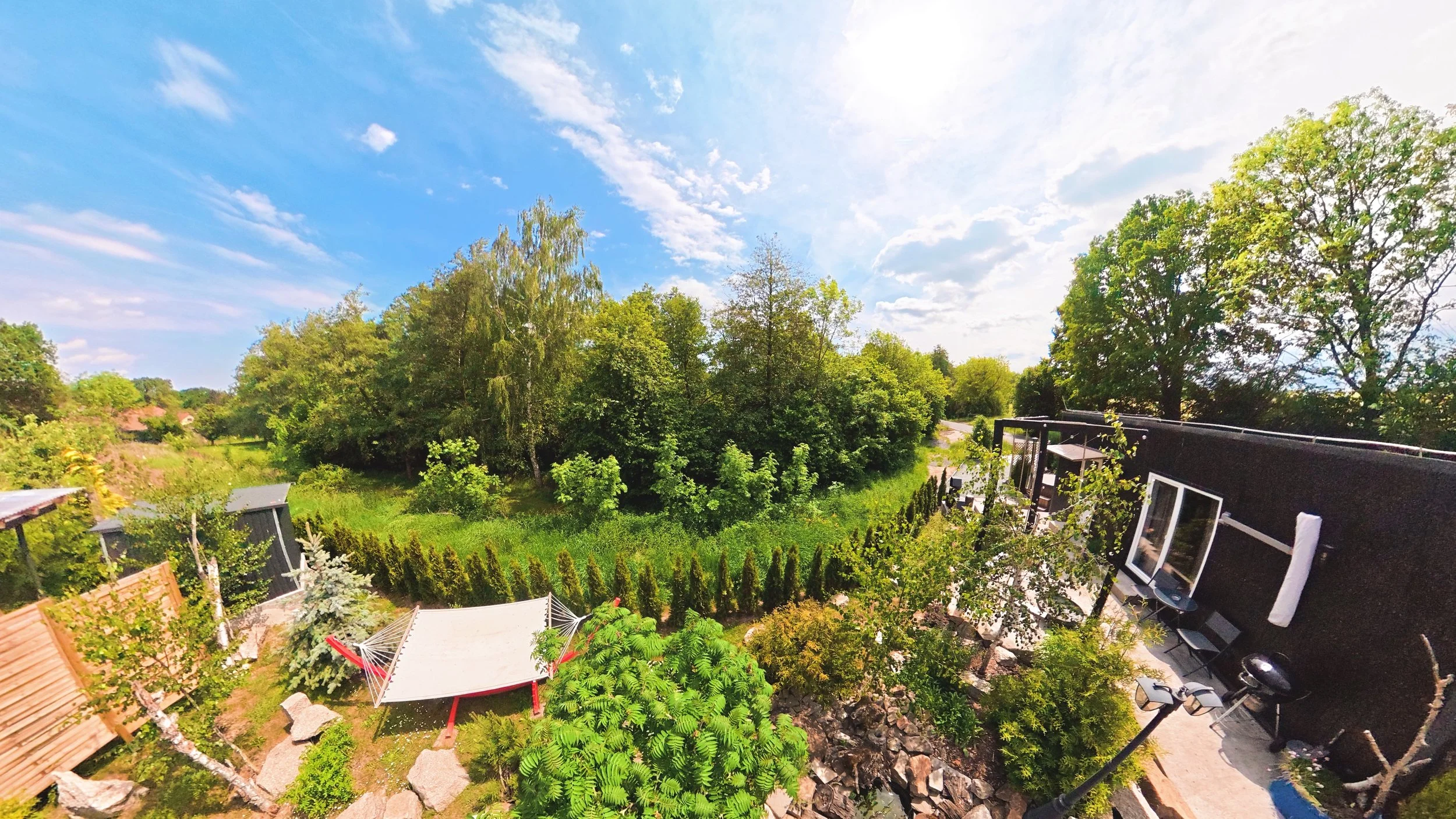 View of a backyard garden with lush green trees, shrubs, a small shelter with a white canopy, and a patio area with outdoor furniture, under a partly cloudy sky.