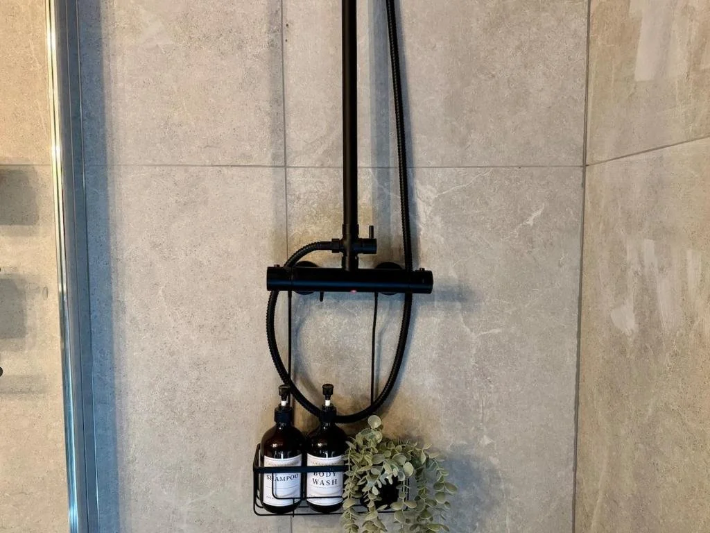 Black shower head with two bottles of shampoo and body wash, and a small bush-shaped plant, mounted on a tiled shower wall.