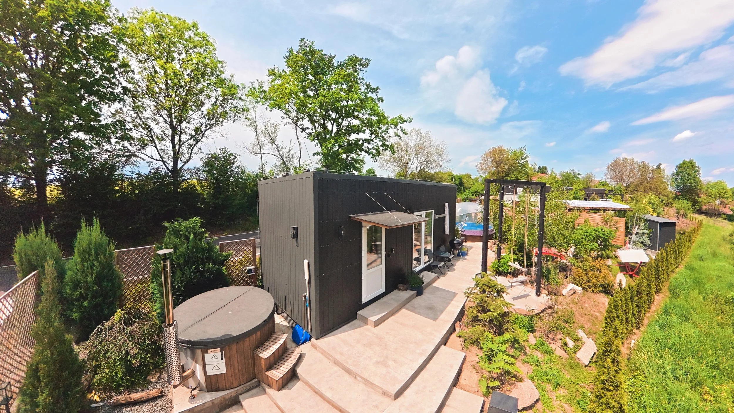 A backyard patio area with a small black and gray shed, hot tub, and outdoor furniture, surrounded by lush green trees under a blue sky with scattered clouds.