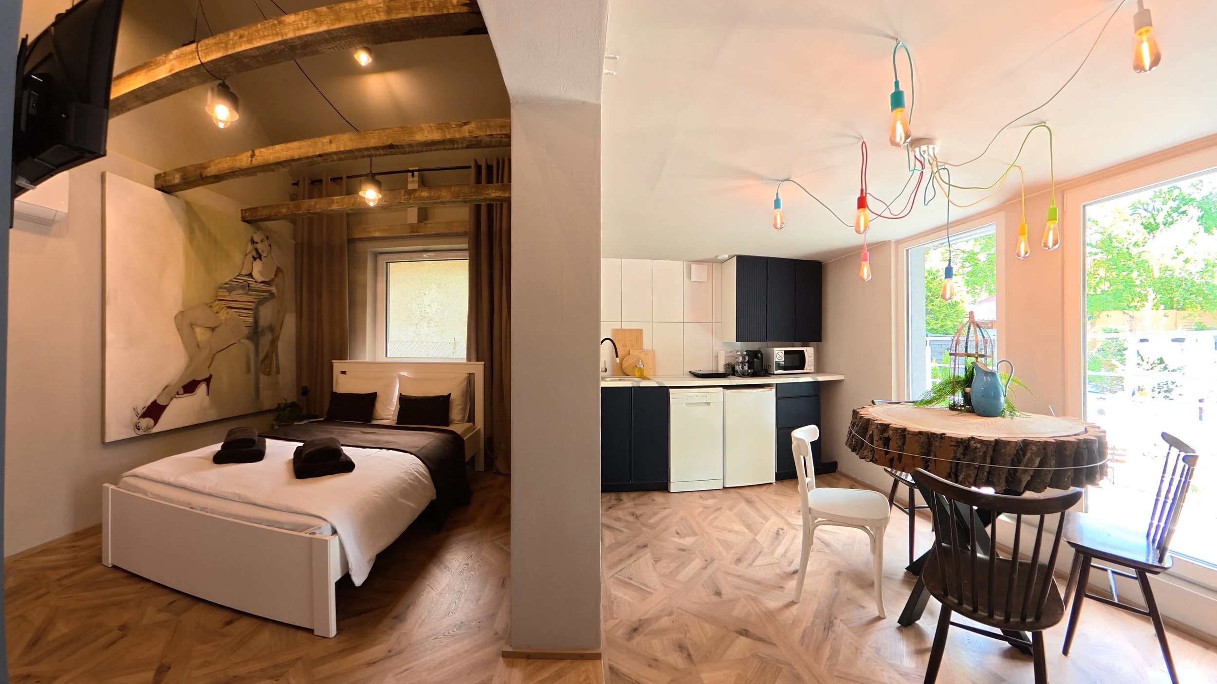 A small apartment with a bedroom and kitchen separated by a partial wall. The bedroom has a bed with black pillows and a large artwork on the wall. The kitchen area has black cabinets, small appliances, and a window with green trees outside. A round table made from a tree trunk has a blue vase with plants and a colorful light fixture hanging from the ceiling.