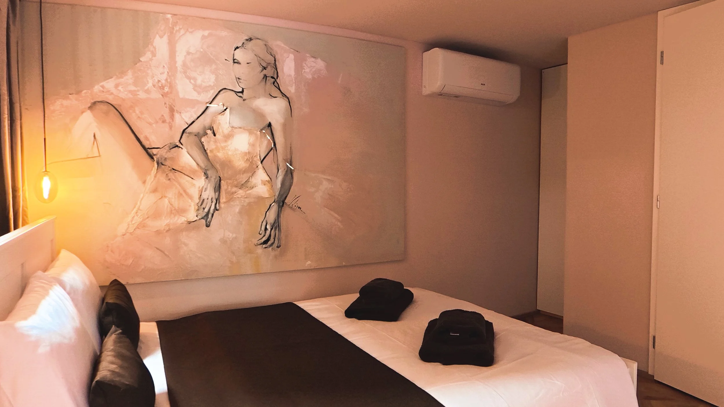A bedroom with a large abstract portrait painting of a woman, a bed with white and black linens, and a wall-mounted air conditioner.