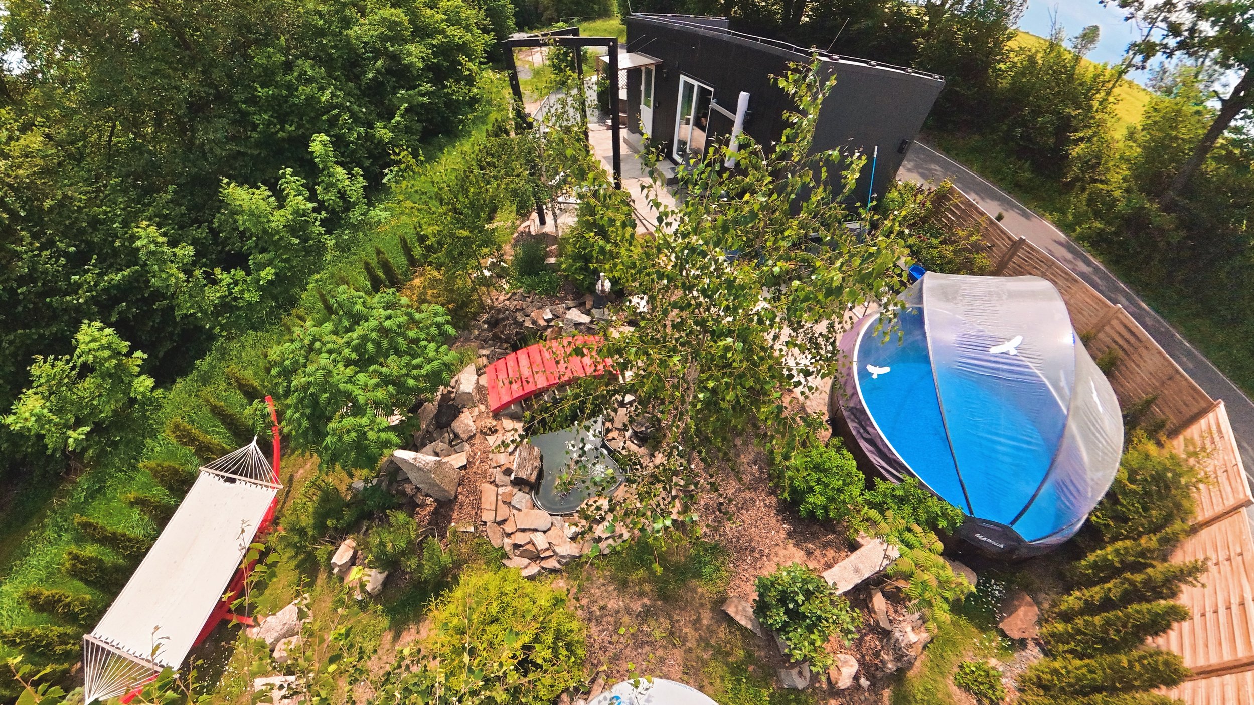An aerial view of a backyard garden with lush green trees, a small pond with rocks, a slide, and a canopy with a seating area. There is a black building and a fenced perimeter visible.