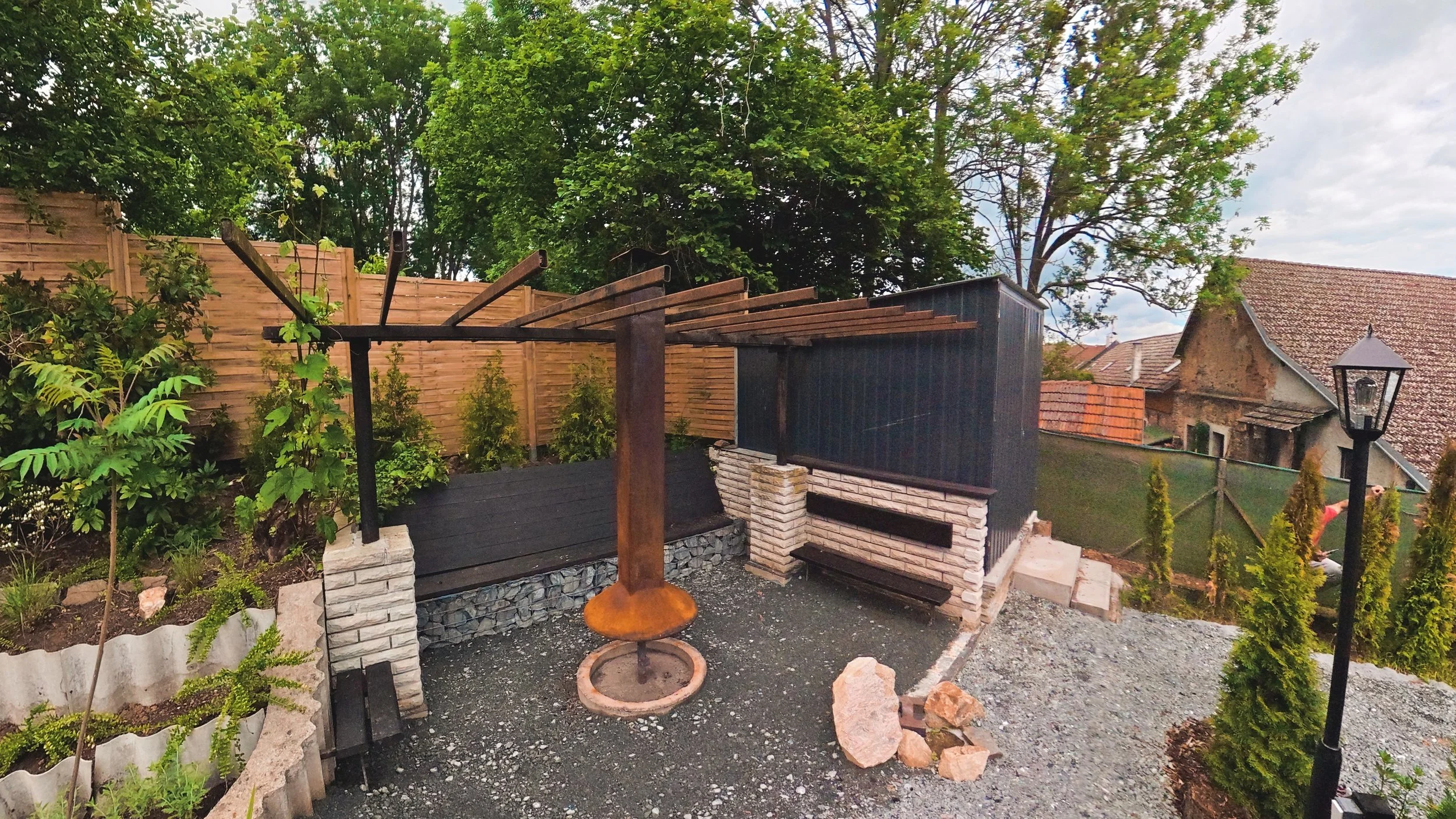 A backyard garden area with a wooden privacy fence, small trees and plants, a black and brown wooden structure, and outdoor lighting fixtures.