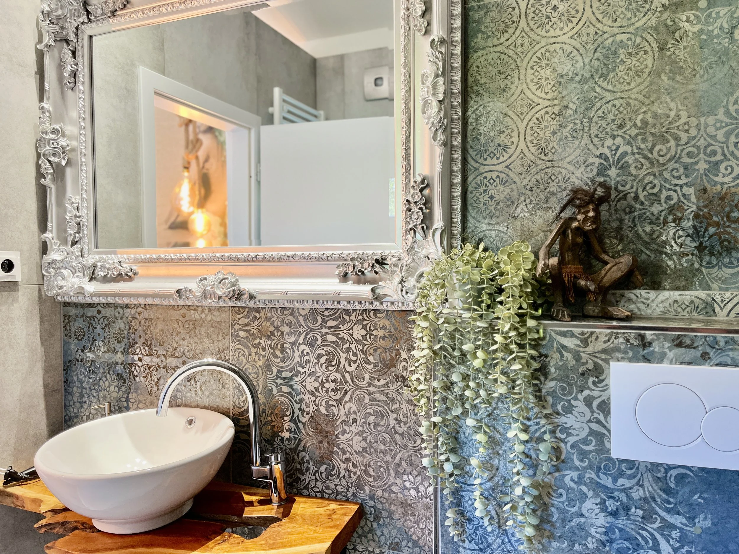Decorative mirror above a bathroom sink with a wood countertop, a small potted plant, and a bronze sculpture of a woman.
