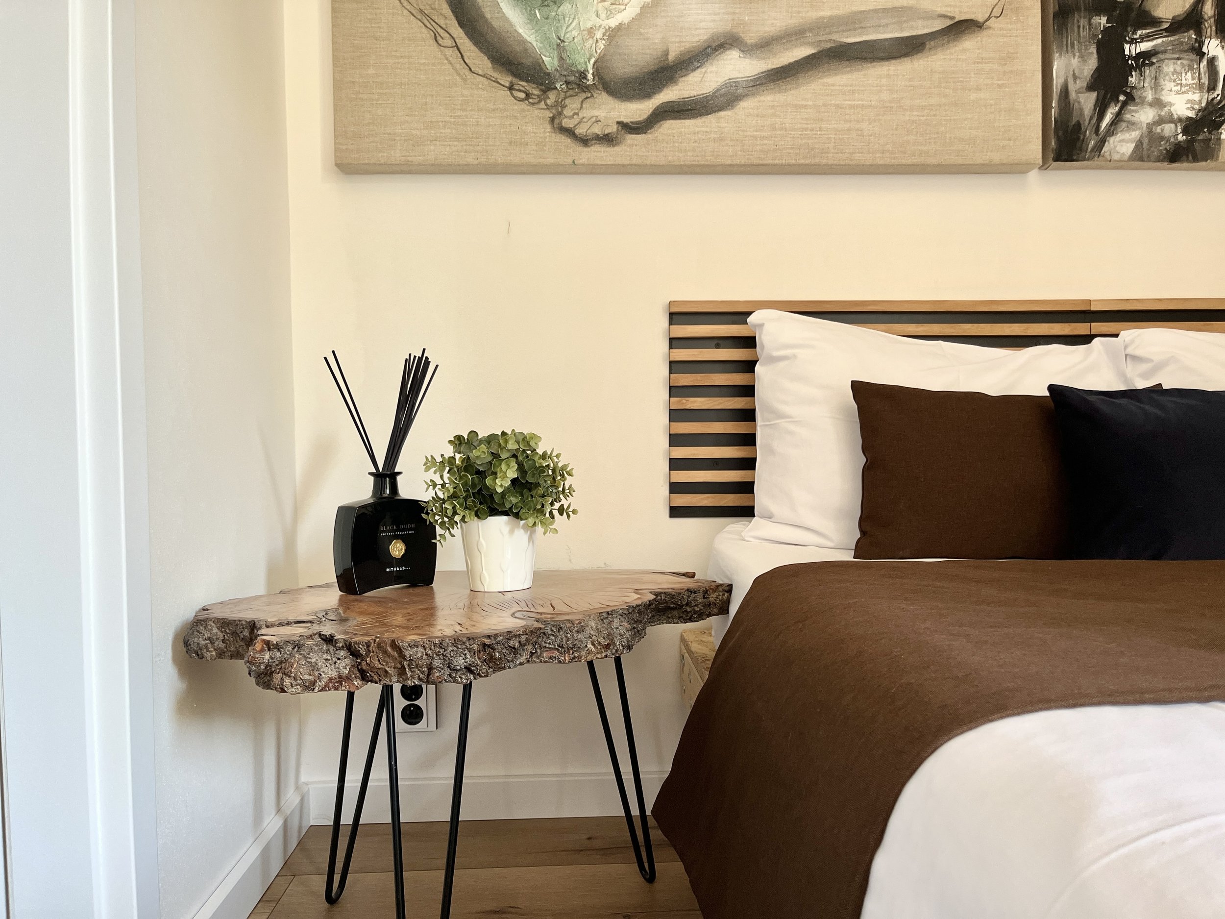 A bedroom corner with a wooden side table holding a black reed diffuser, a potted succulent, and a white vase. Part of a bed with white and brown pillows and a wooden headboard is visible. Above, a beige canvas painting hangs on the wall.