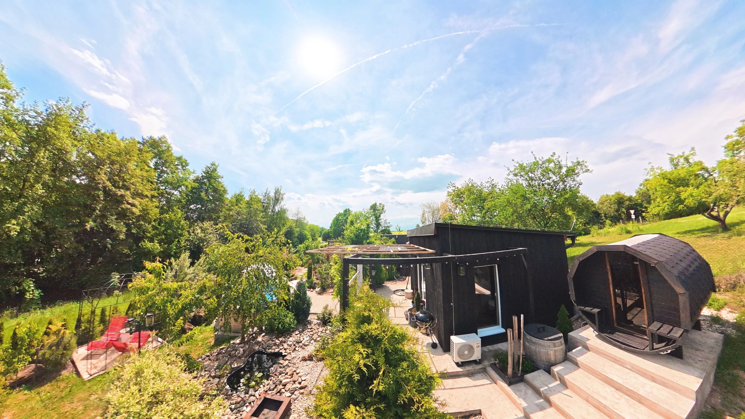 View of a backyard garden area with trees, a black shed, outdoor seating, and a bright blue sky with scattered clouds and the sun.