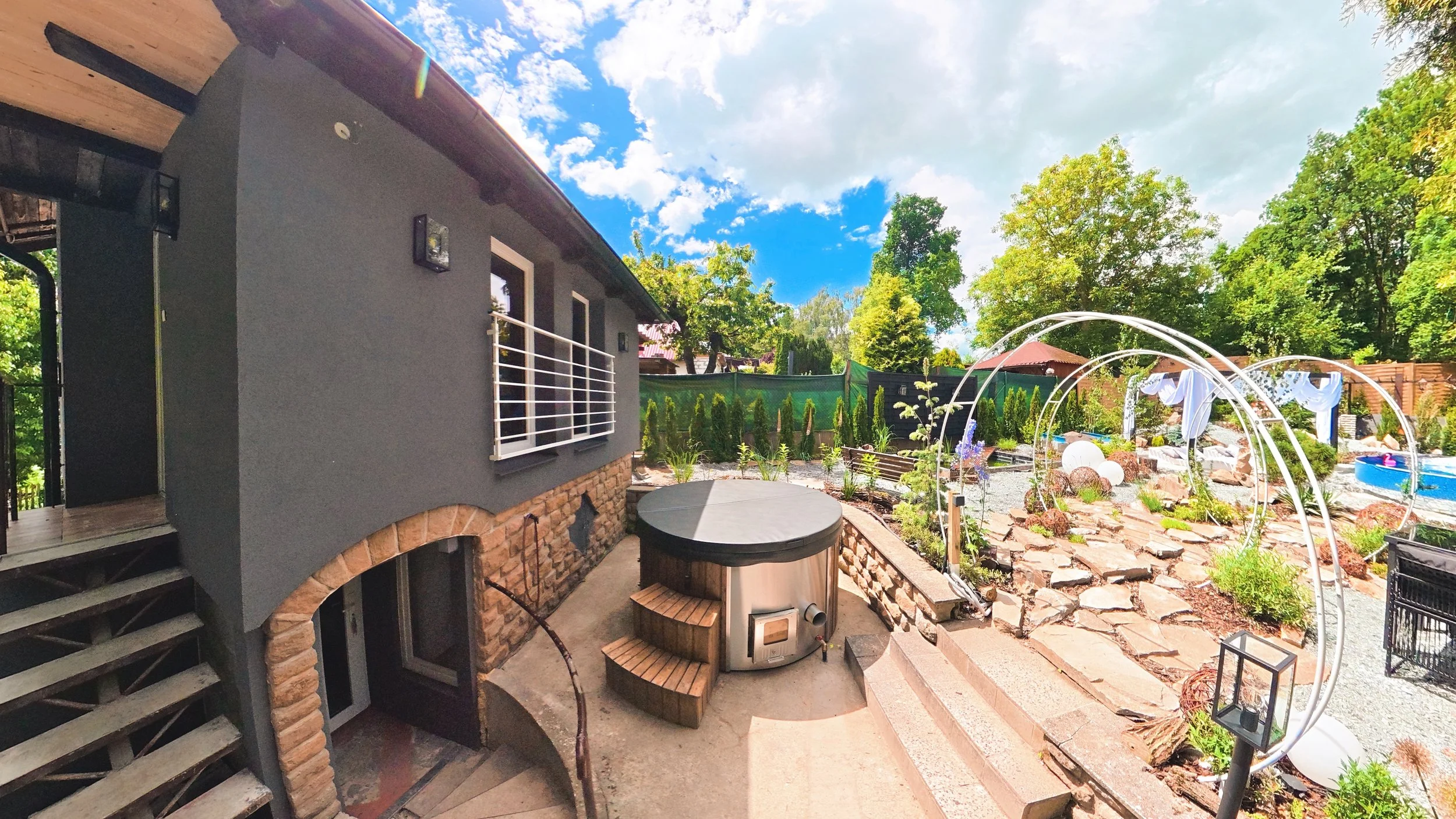 A backyard garden with a gray house on the left, a wooden staircase, and a stone patio. The garden features lush green trees, decorative arches, and outdoor seating with drapes. The sky is partly cloudy with blue sky visible.