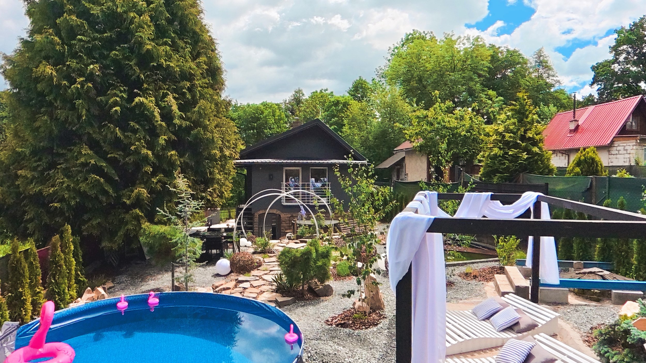 A backyard with tall green trees, a small dark gray shed, and a neighboring house with a red roof. There is a small pool with pink pool toys, a garden area with stepping stones, and a clothesline with white fabric hanging on a wooden frame.