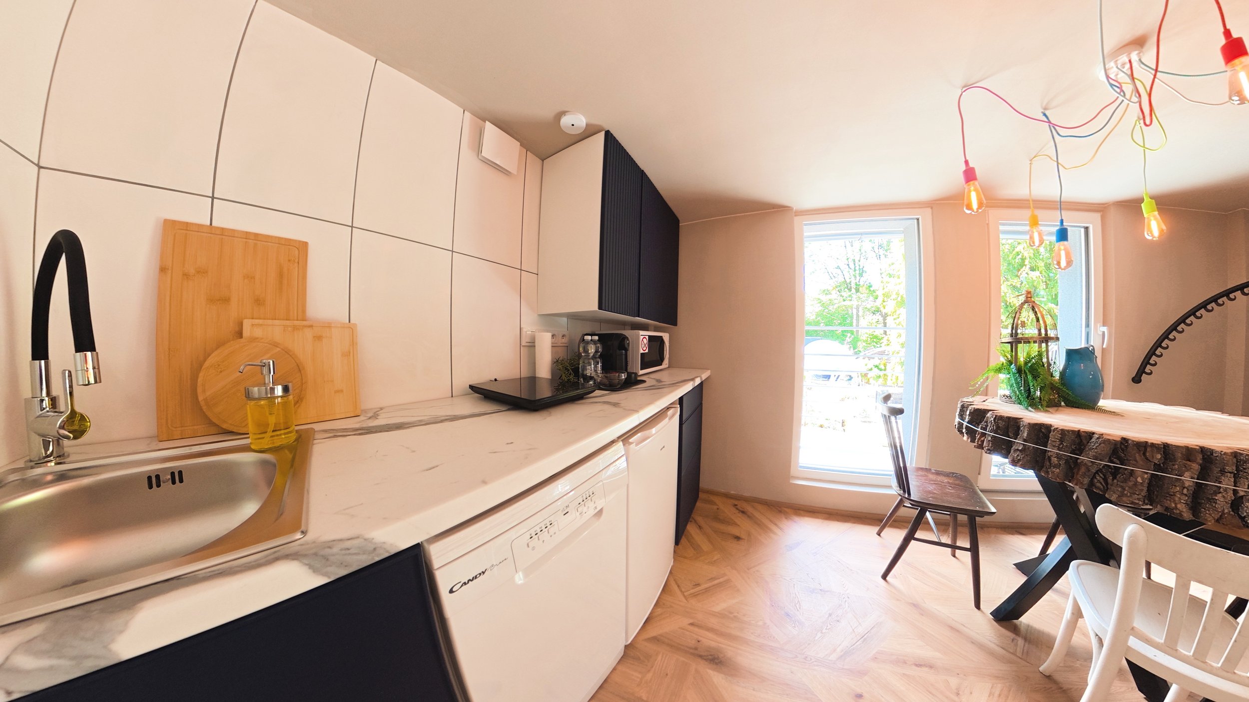 Bright kitchen with white tiled walls, marble countertop, black cabinets, sink with yellow soap, cutting boards, coffee maker, microwave, window with greenery outside, wooden tree slice dining table with decorative jars and plants, colorful hanging light bulbs, and a white chair.