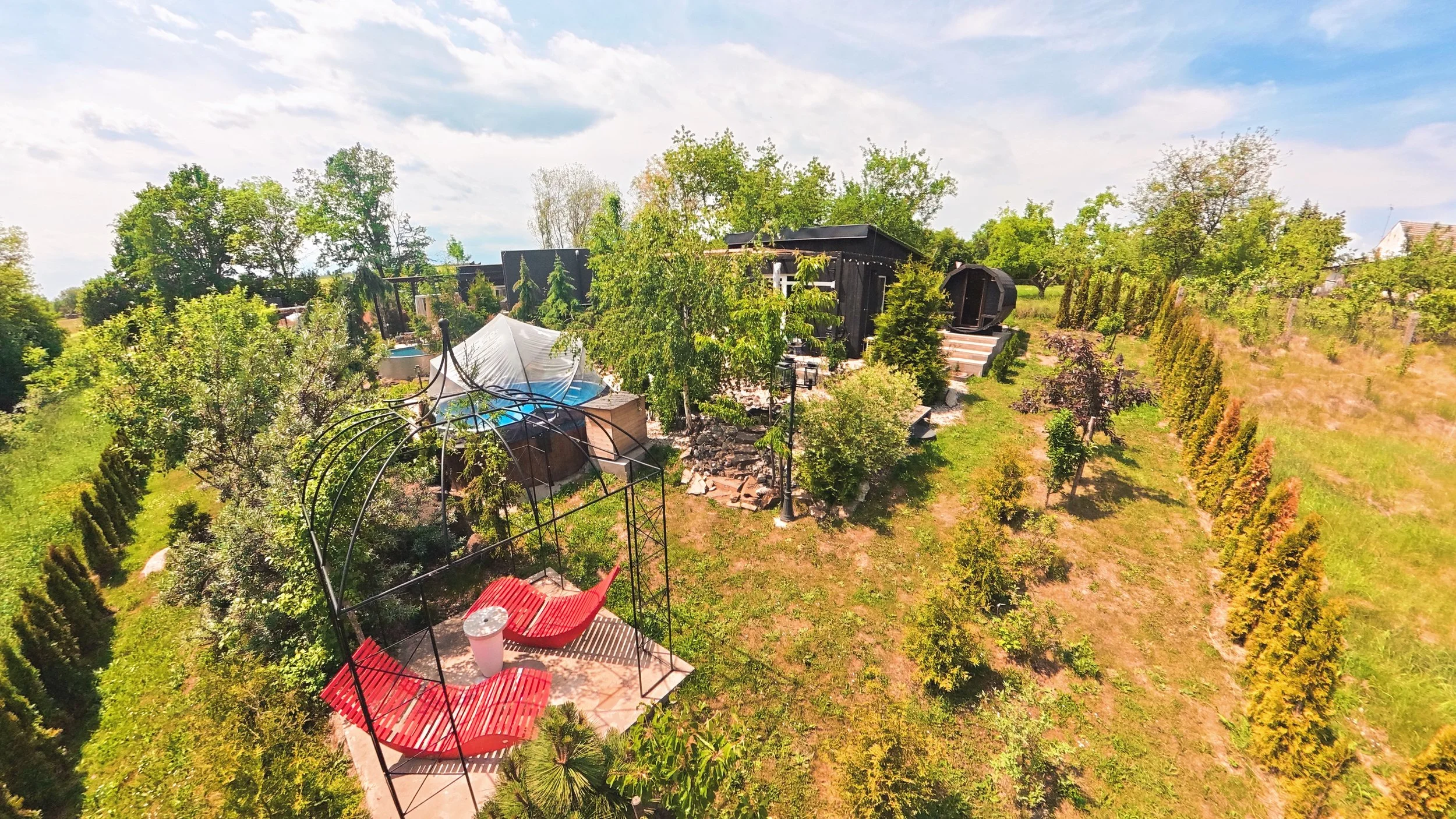 Aerial view of a lush backyard with trees, a small patio with red chairs and a table, a greenhouse, and a black shed, under a partly cloudy sky.