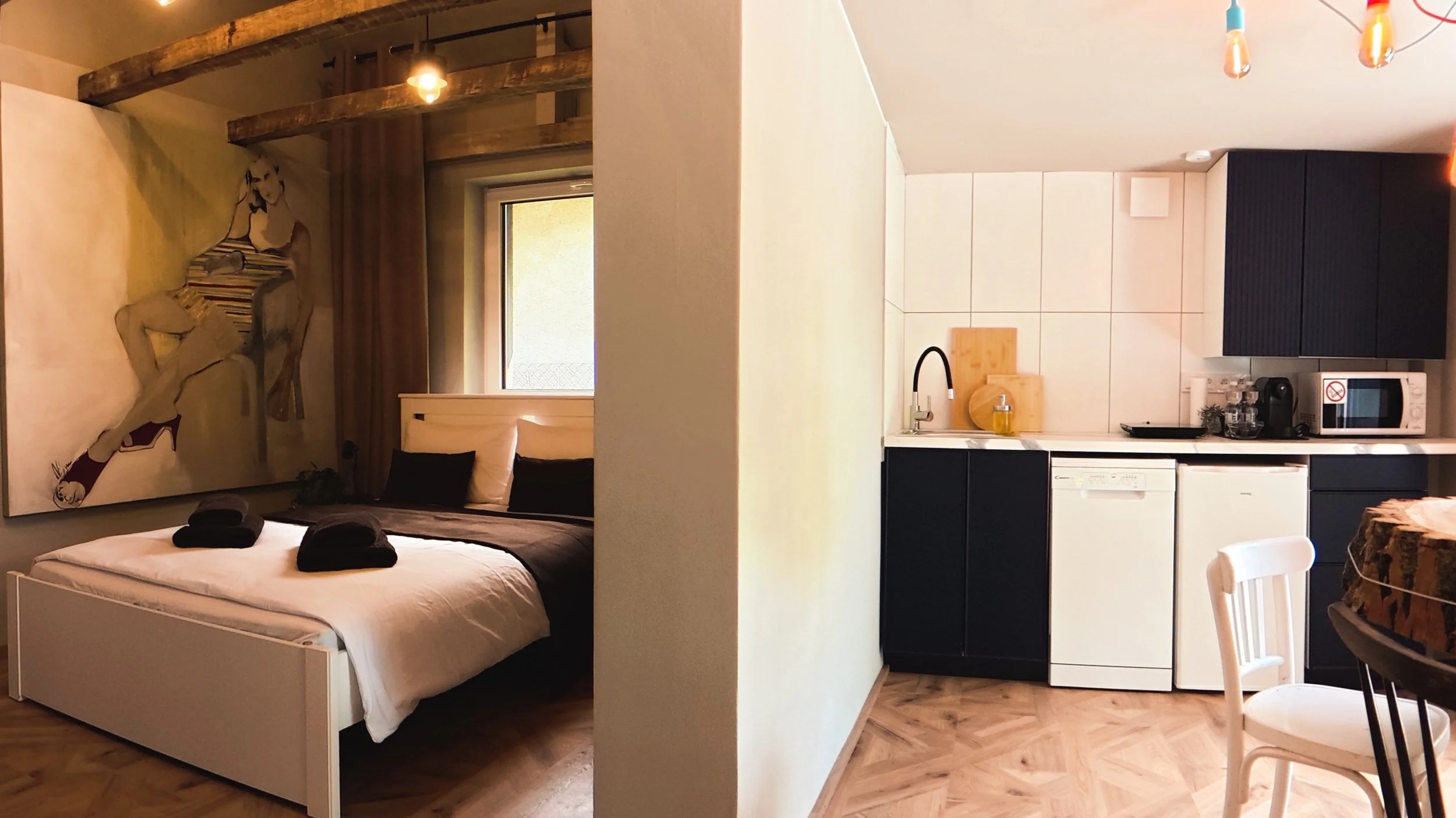 A cozy bedroom with a large bed, dark bedding, and artwork of a woman on the wall. The room has warm lighting and wooden accents, with a window behind the bed. Adjacent is a compact kitchen with black cabinets, white backsplash, and small appliances, including a microwave and coffee maker, with a table and chair nearby.