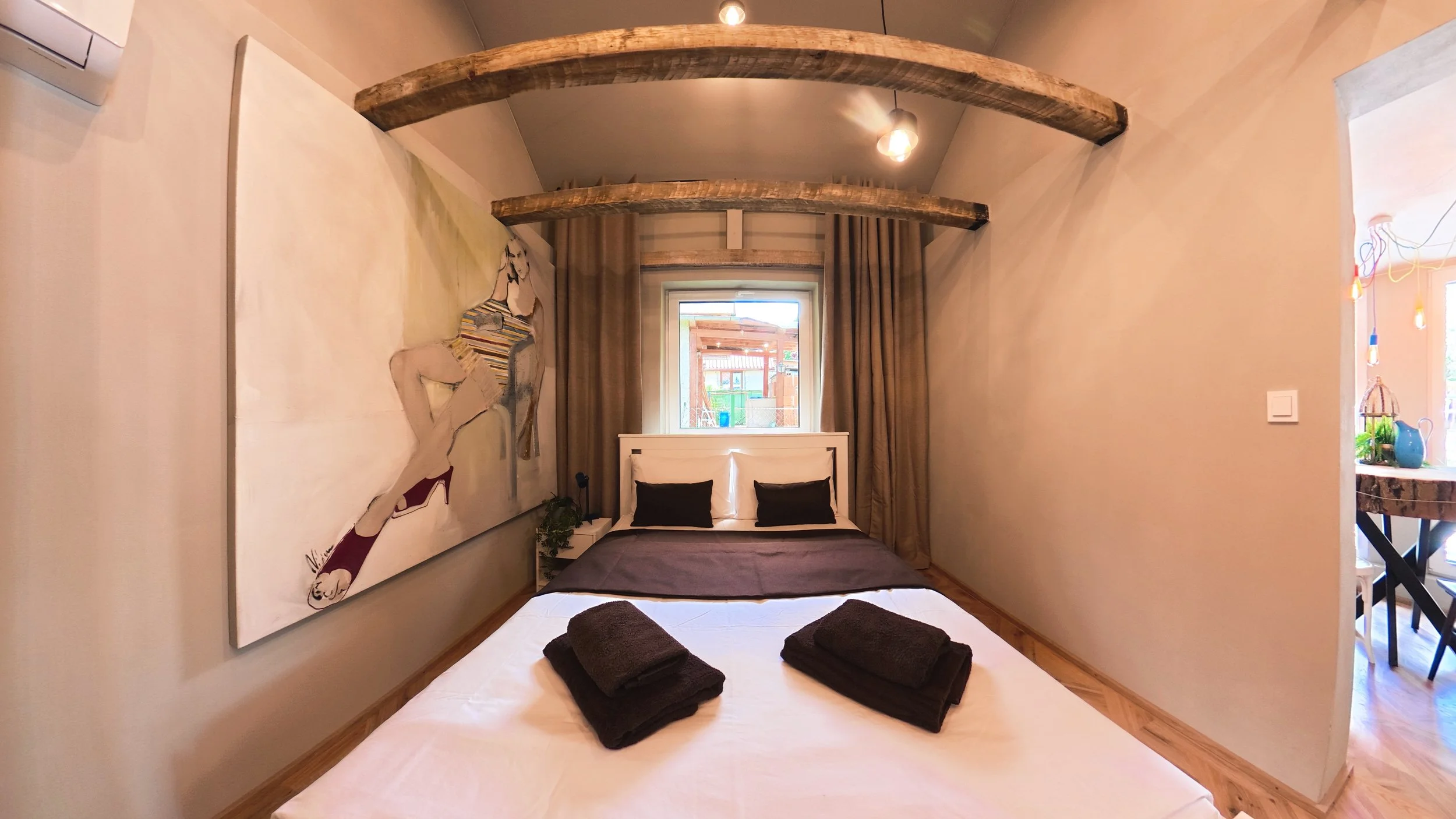 A bedroom with a white bed, black pillows, and folded dark towels on top. On the wall to the left, there is a large painting of a woman in striped clothing and heels. The room has wooden beams on the ceiling, a window behind the bed, and beige curtains. To the right, part of an open area with decorative hanging lights and a table with plants and vases is visible.