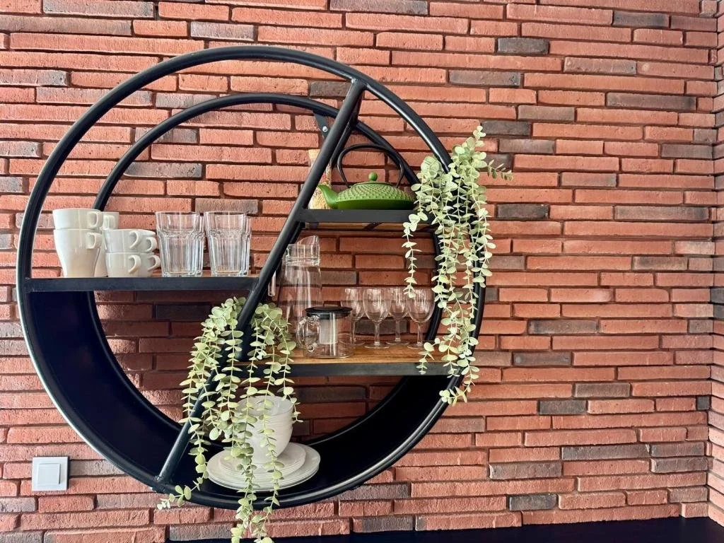 A round black wall shelf with three compartments, mounted on a red brick wall. The shelf holds white cups and glasses, a green teapot, glass jars, plates, wine glasses, and decorative hanging greenery.