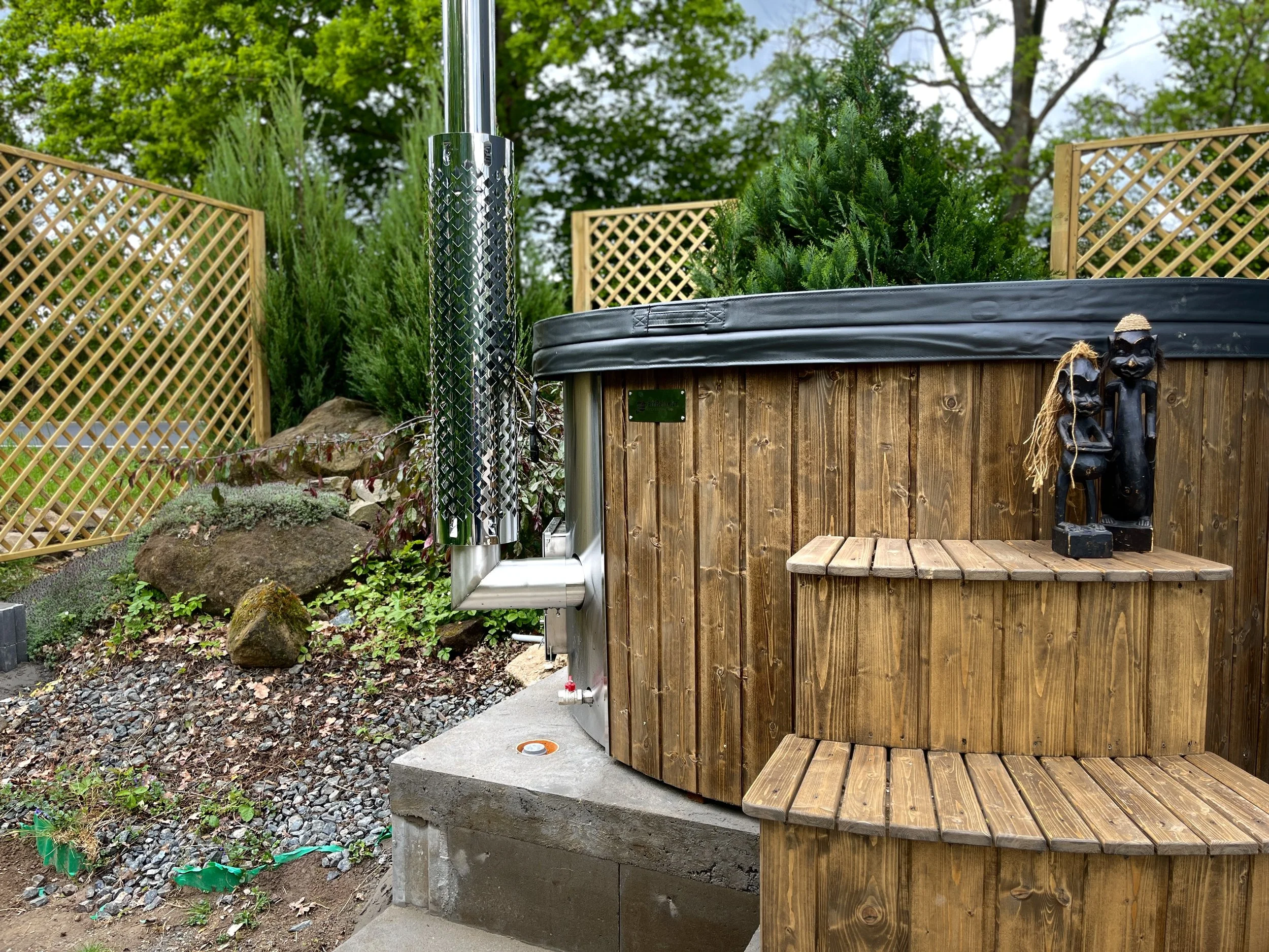 An outdoor hot tub made of wood with a cover, situated on a concrete base. To the right of the hot tub, there are two African-style sculptures and a wooden bench. In the background, there are trees, bushes, and a lattice fence, with a clear sky above.