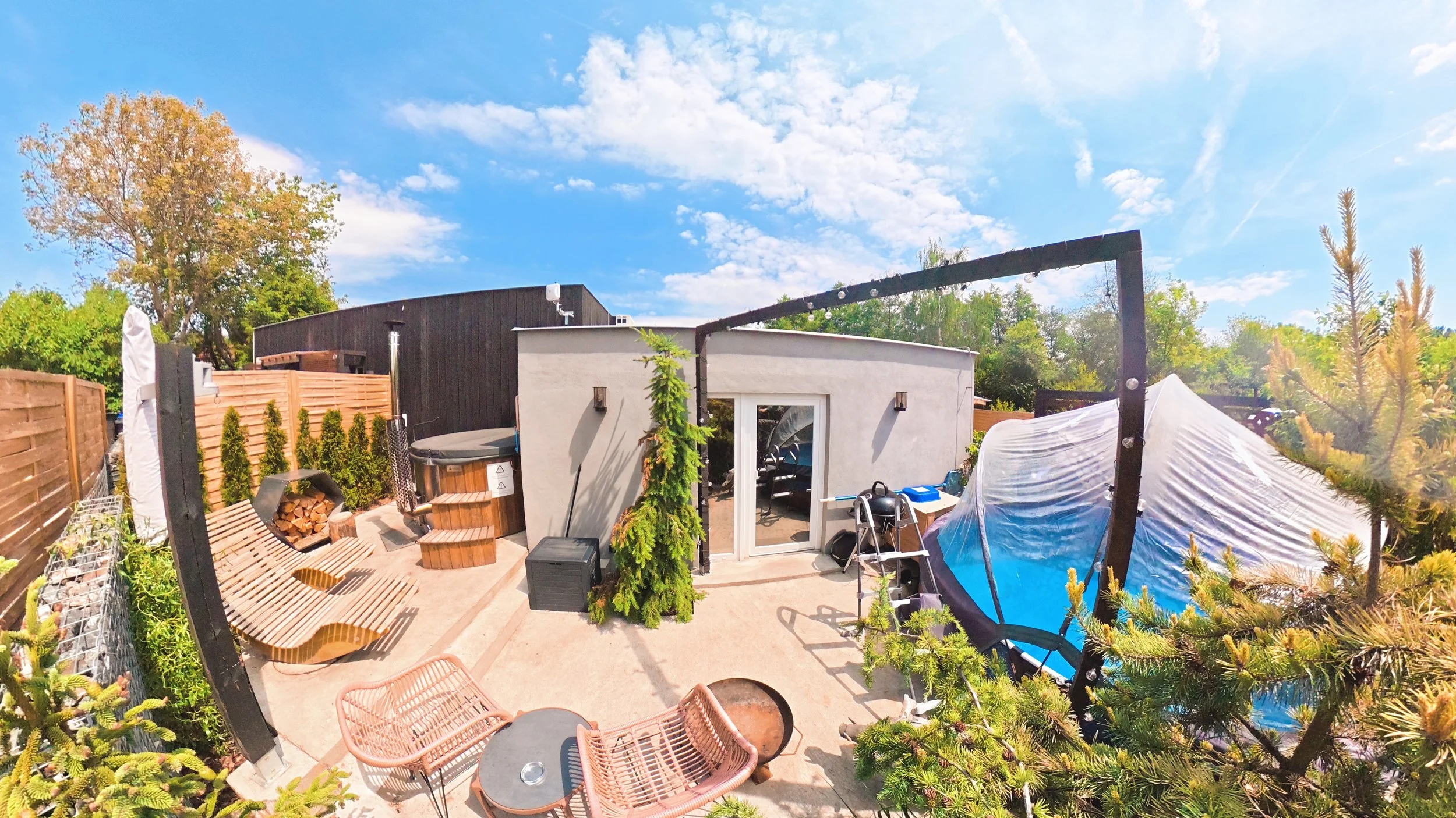 A backyard patio area with outdoor furniture, a hot tub, small shed, and garden plants, under a bright blue sky with clouds.