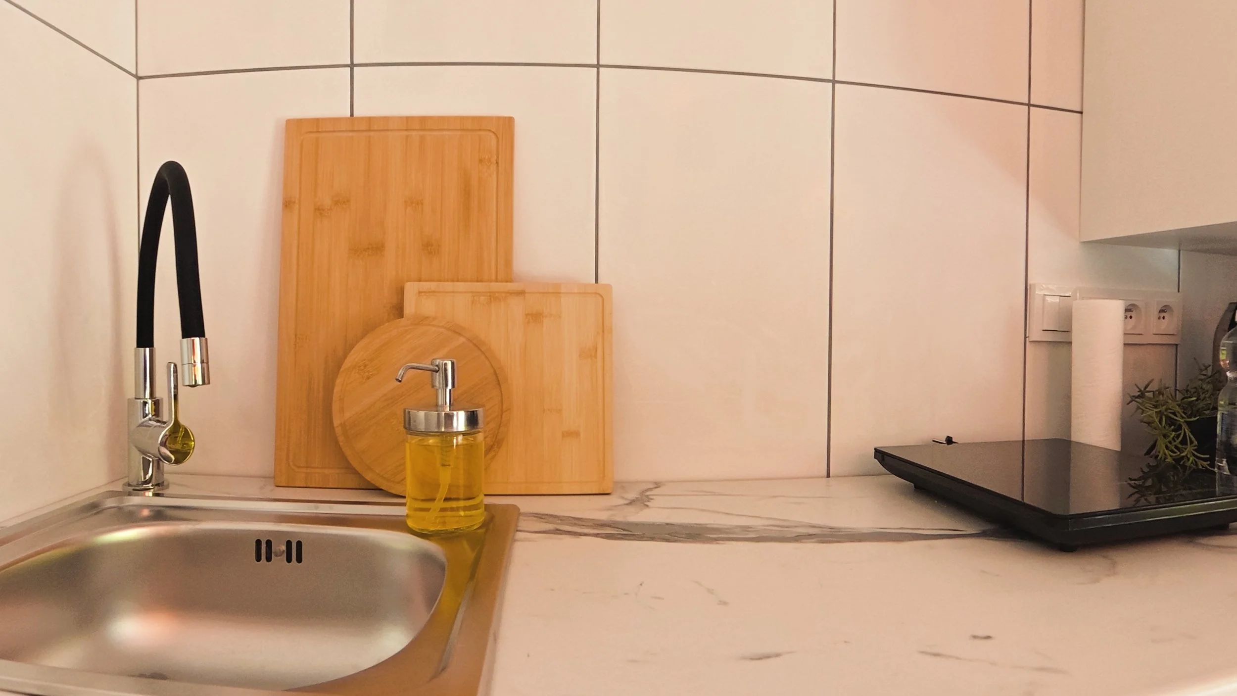 Kitchen counter with a sink and faucet, a bottle of yellow dish soap, wooden cutting boards, a small plant, a portable stove, and an electrical outlet on the wall.