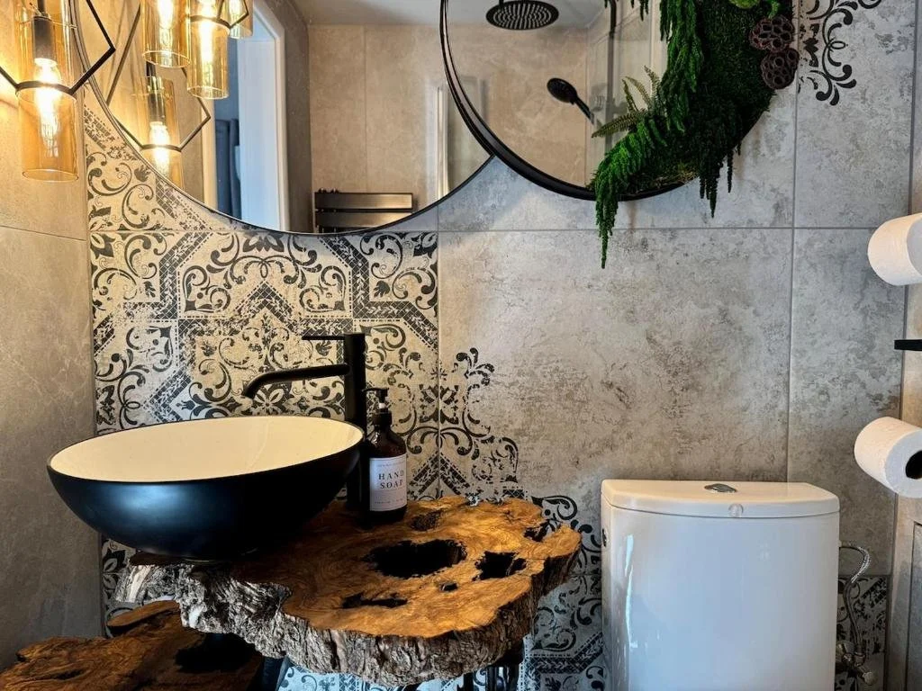 Bathroom with black-and-white patterned tile wall, wooden sink console, black vessel sink, black faucet, large round mirror, decorative wall lamp, soap bottle, white toilet, and wall-mounted toilet paper dispenser.