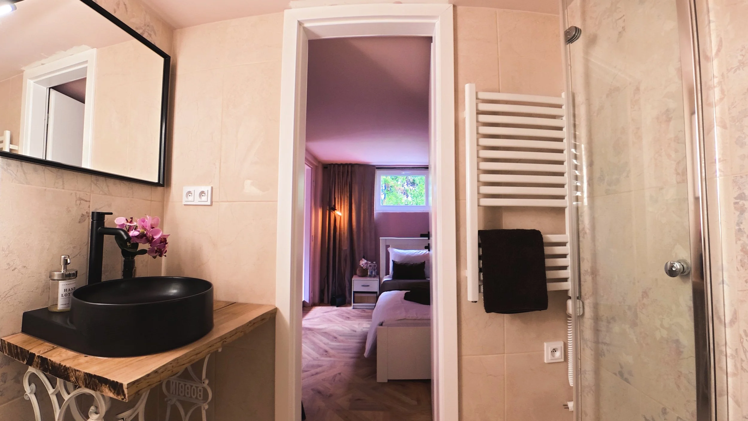 Bathroom with a black sink, a mirror, a white towel warmer, and a view into a bedroom with a bed, window, and wooden floor.