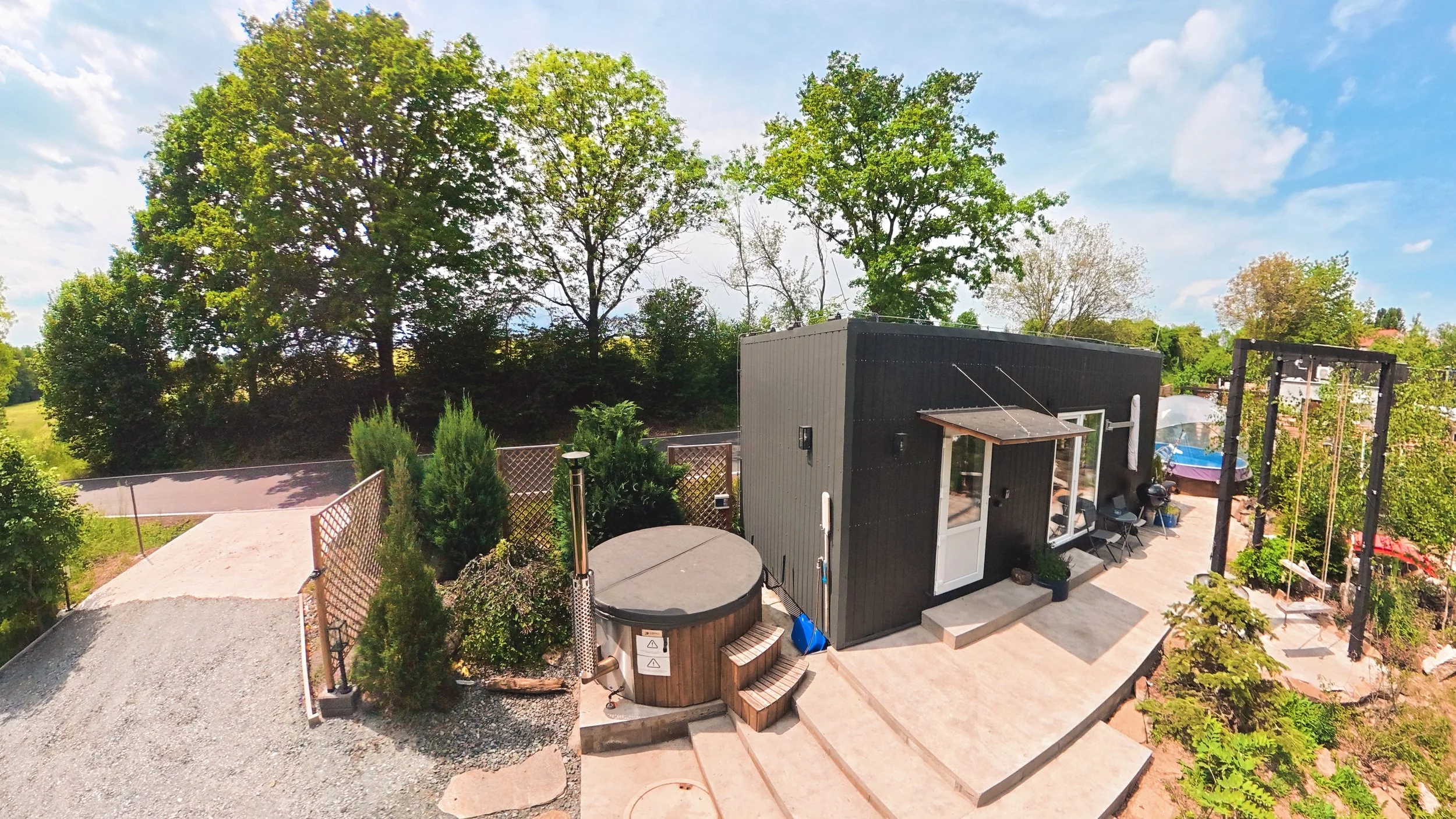 A backyard with a small modern gray shed, a hot tub, a patio, and lush green trees under a partly cloudy sky.